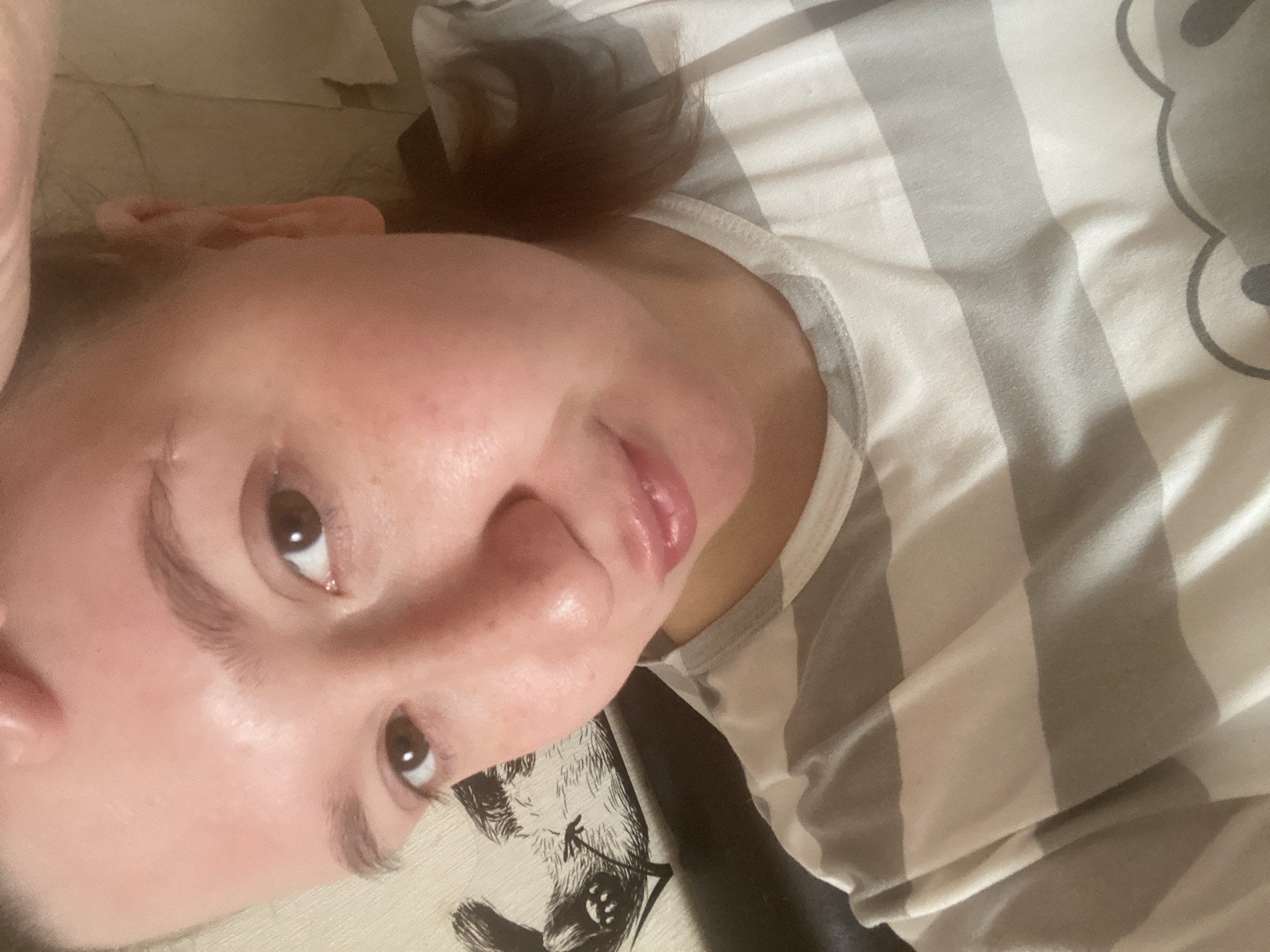 A person is lying on their back, close to the camera. The person has light skin, brown eyes, and brown hair pulled back. They are wearing a striped white and gray shirt. Beneath the person appears to be a pillow with a design of an animal face. The scene is indoors. The city of Chaikovsky, Russia, is not discernible from the image.