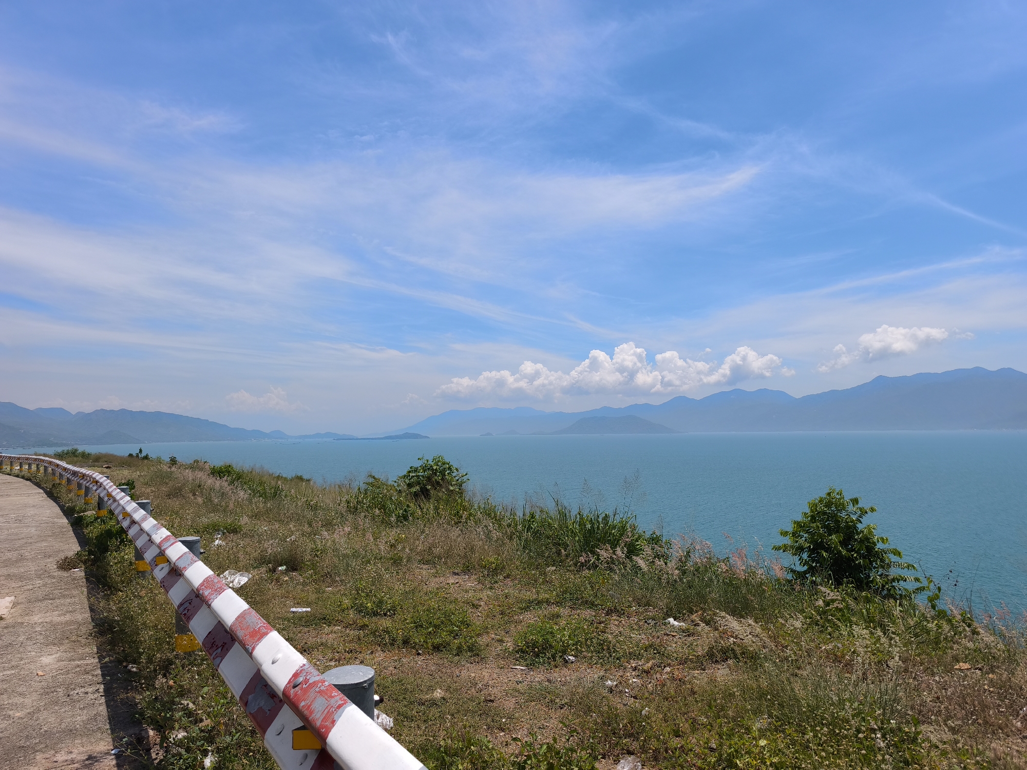 A scenic view of the ocean with mountains in the distance is captured from a paved road with a red and white striped guardrail. The sky is a clear blue with scattered white clouds. The road curves to the left, bordered by dry grass and a few small trees. The water is a vibrant turquoise, and the mountains in the background are a hazy blue. The overall impression is one of a peaceful coastal landscape. The image was taken in the city of Bac Nha Trang Ward, Vietnam.