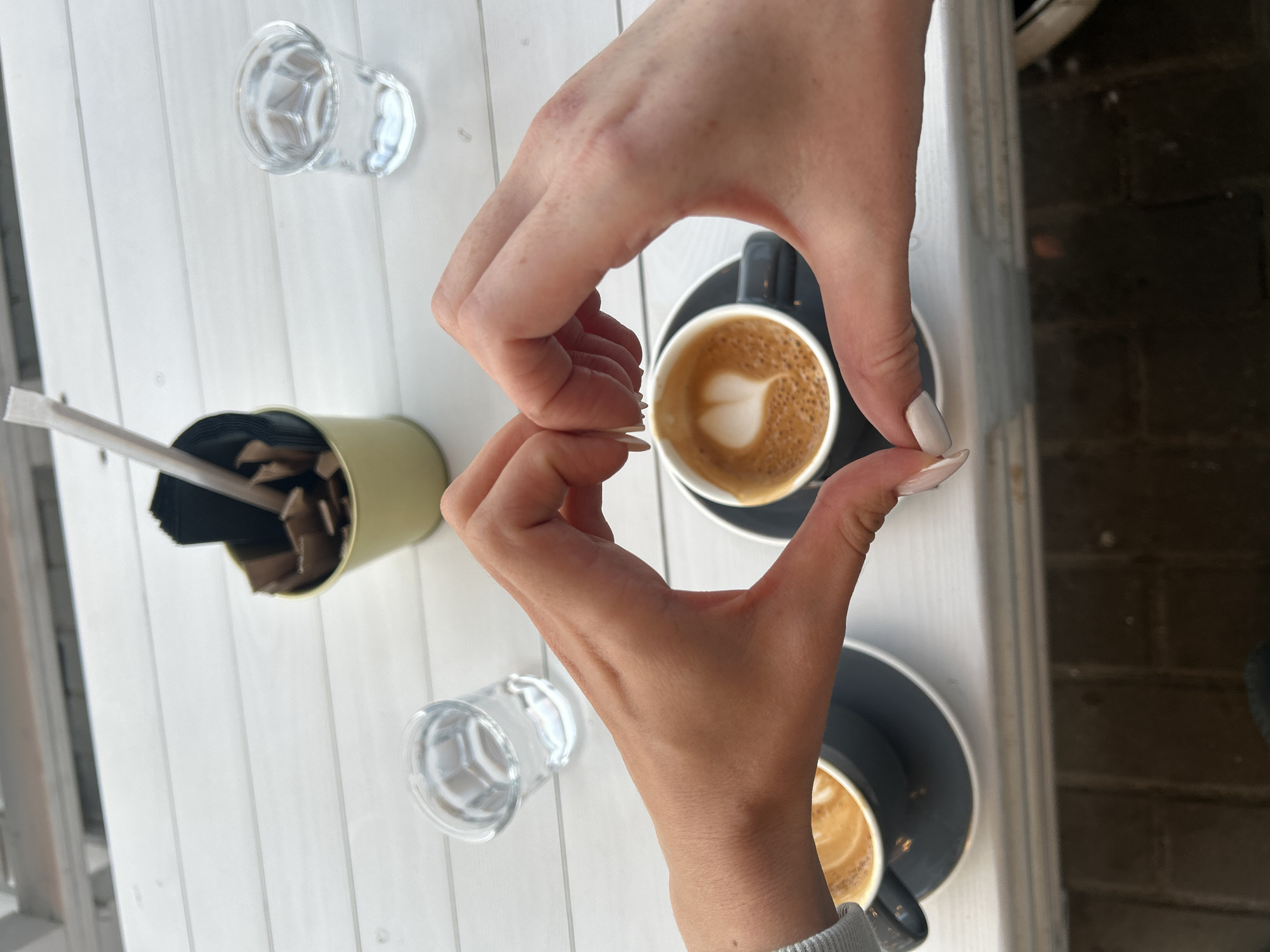 A top-down shot captures two hands forming a heart shape over two cups of coffee on a white wooden slatted table in Tolyatti, Russia. The hands, appearing to belong to a woman with white nail polish, frame one of the coffee cups, which features a latte art heart in its foam. A second identical coffee cup, also with latte art, is visible further down on the table. Both coffees are served in dark grey or black ceramic cups with matching saucers.
Alongside the coffee, two small, clear glass tumblers are present, one near each coffee cup. A light olive-green cylindrical container, possibly a caddy for sugar packets or stirrers, is positioned in the upper left, with a white straw partially visible sticking out. The contents of the container appear to be individually wrapped sugar packets or stirrers.
The bright lighting suggests it is daytime. The setting appears to be an outdoor cafe or patio, indicated by the white wooden table and a dark, textured background on the right side of the frame, possibly brickwork or pavement. The overall mood conveyed by the heart gesture and two coffees is one of affection, companionship, and enjoyment. There is no visible text in the image.