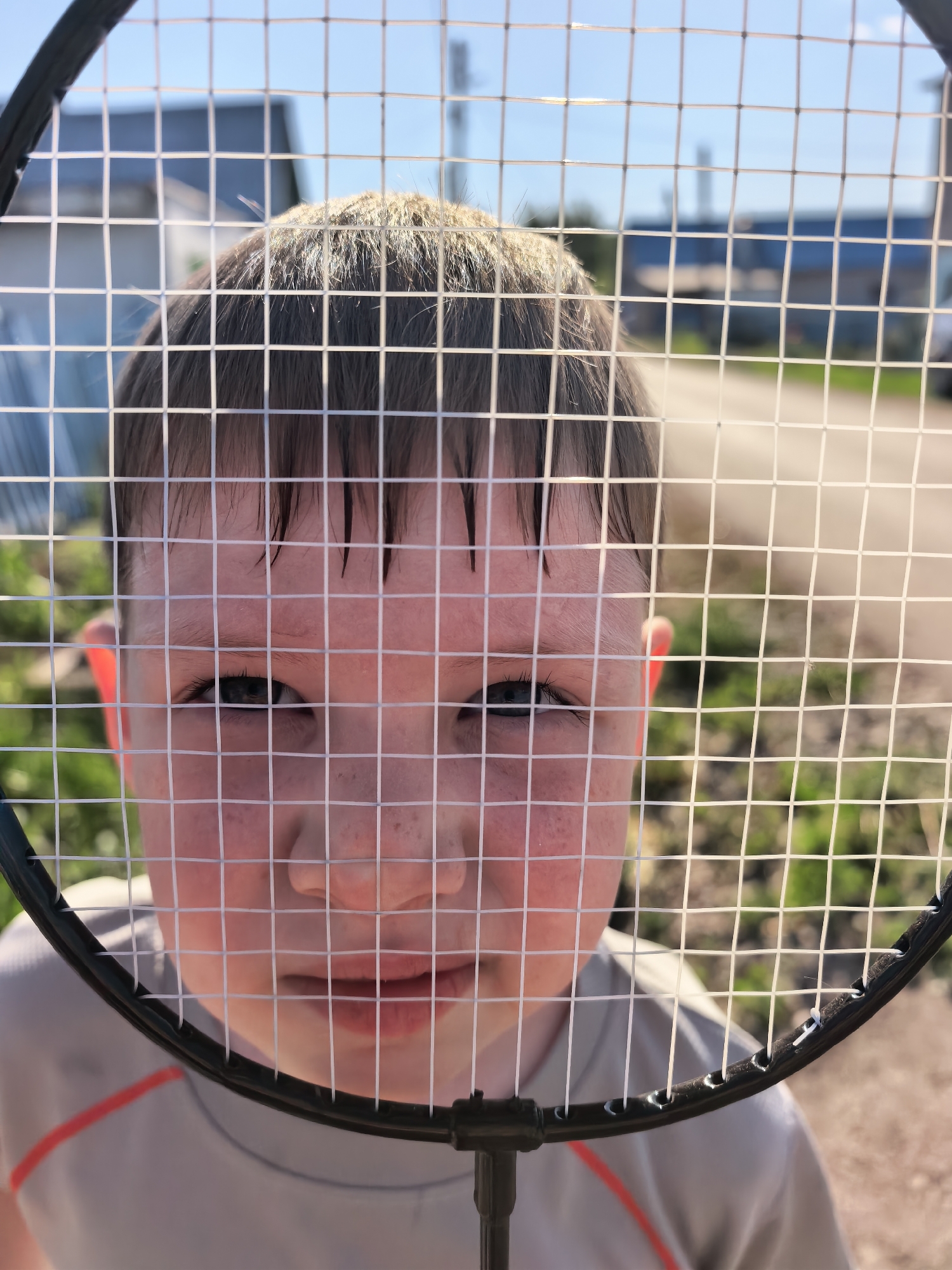 A close-up, portrait-style outdoor shot captures a child's face through the mesh of a badminton racket. The child has fair skin, brown hair, and is wearing a light grey shirt with an orange accent. The background reveals a blurry exterior scene with buildings and foliage under a bright, blue, cloudless sky. The specific location is presumed to be within Sterlitamak, Russia.