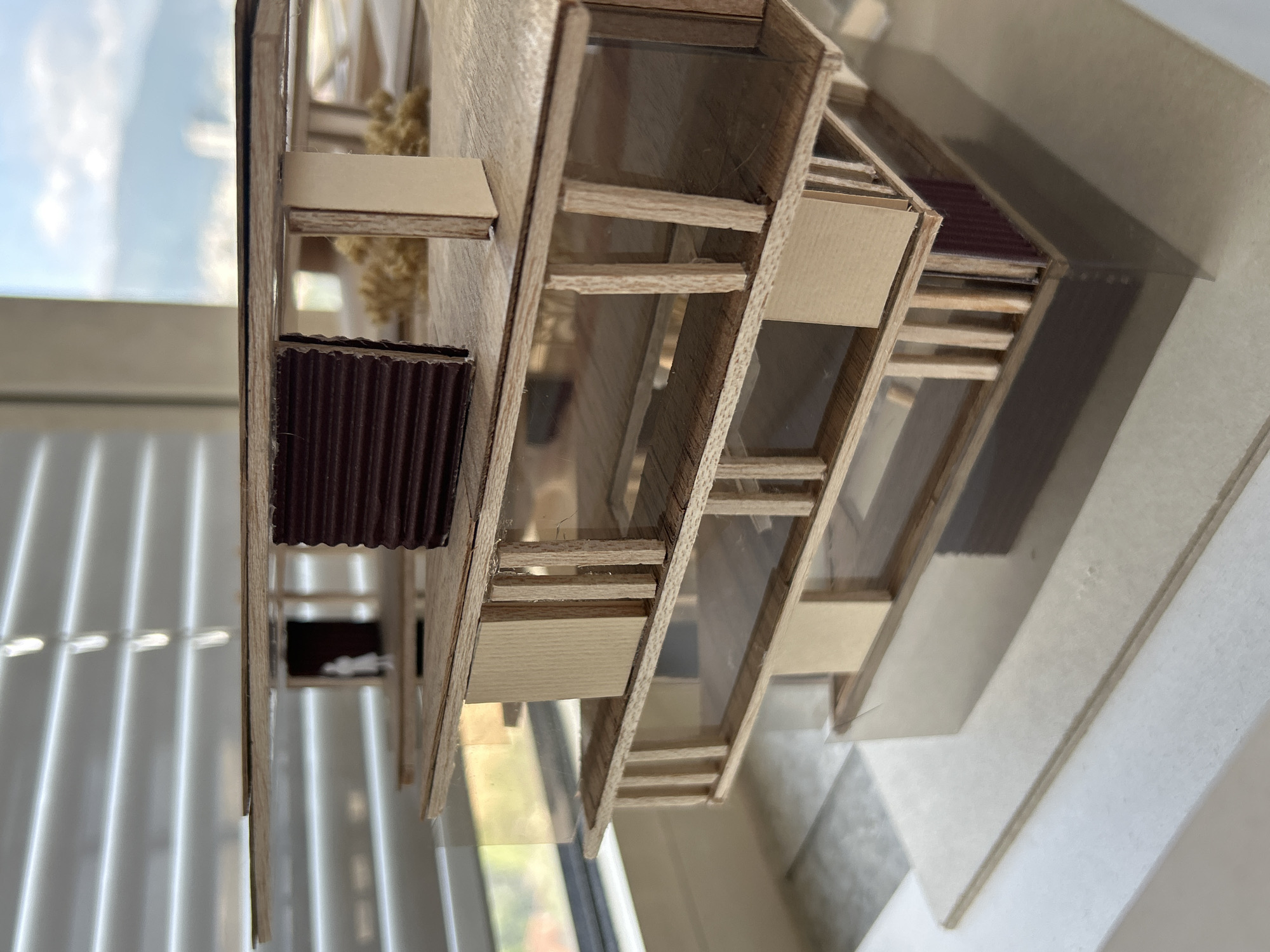 The image shows an architectural model. The model appears to be made of wood and cardboard. It consists of several levels and features details like windows, a corrugated roof element, and support beams. The model is placed on a white surface, possibly a table. The background includes a window with venetian blinds and a view of a blue sky and green trees. There is no evidence to identify the building is in Sofia, Bulgaria.