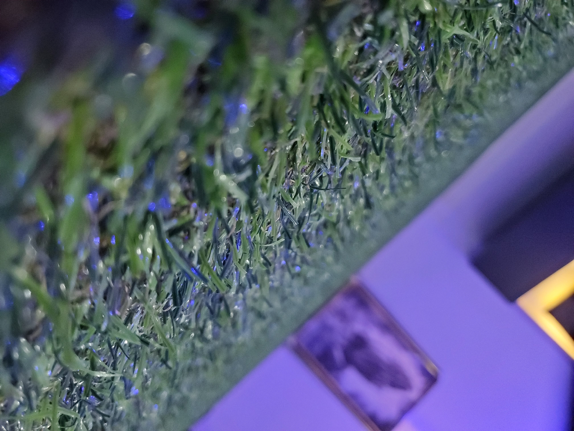 The image is a close-up, low-angle shot of artificial grass with tiny water droplets or reflective particles scattered across the blades, creating small blue and white highlights. The grass occupies the foreground and extends diagonally upwards from the bottom right to the top left of the frame. In the blurred background, a purple wall is visible, with a framed object with a lighter border hanging on it. To the left of the framed object is a dark object with a yellow accent. The lighting appears to be artificial, with the purple hue suggesting ambient lighting. There are no people, animals, or any other discernable subjects or actions in the image. The scene is devoid of any recognizable landmarks or contextual cues that would indicate a specific location in Bukuru, Nigeria. The overall impression is of a still life composition focusing on texture and light play on artificial grass.