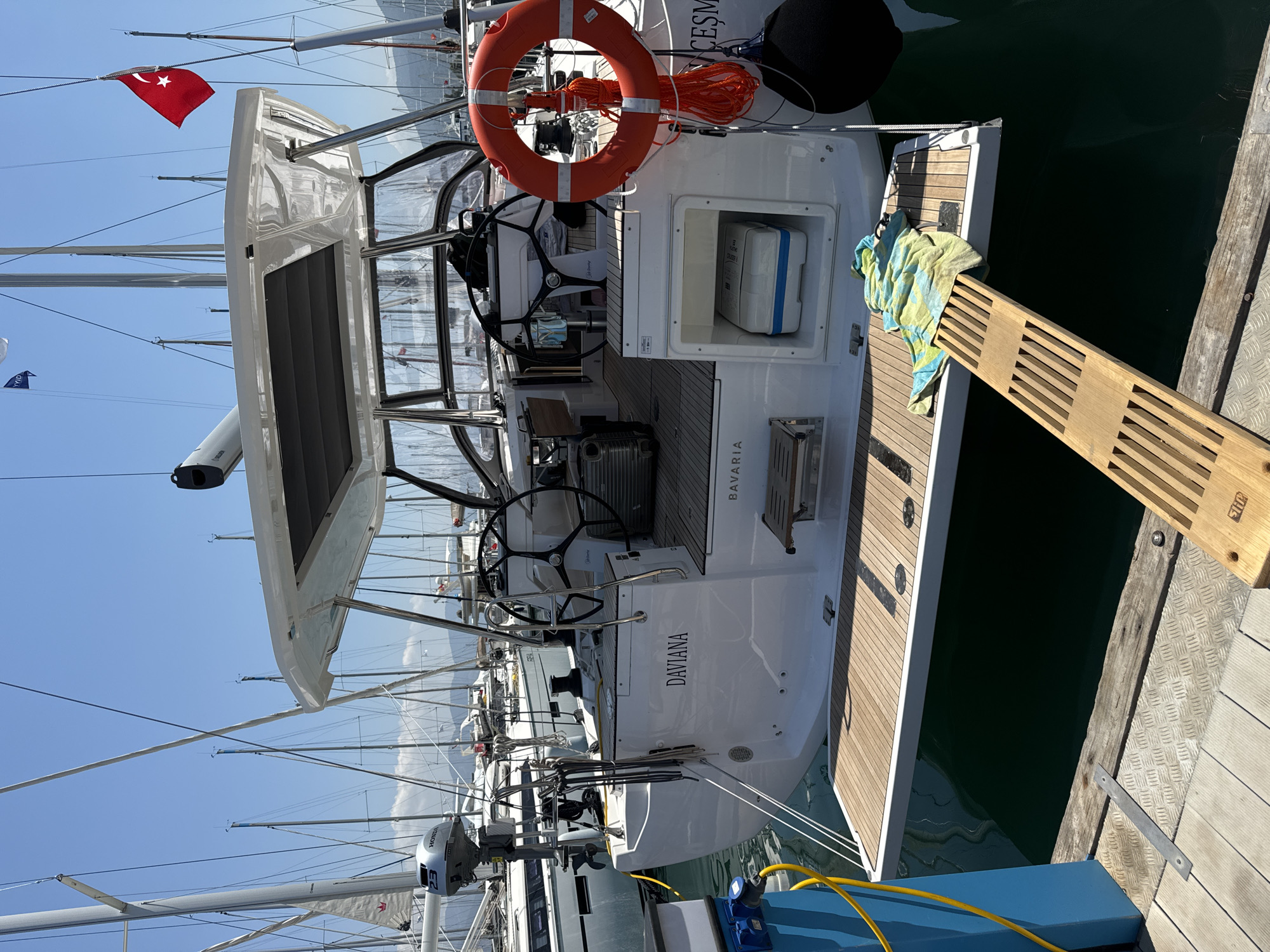The image presents a detailed view of the stern of a white Bavaria sailboat, named "DAVIANA," docked in a marina in Fethiye, Turkey, under a clear, bright blue sky. A red Turkish flag with a white star and crescent is visible, flying from the rigging of an adjacent vessel.
The main sailboat features a spacious teak-decked cockpit with dual steering wheels. A white, hard-sided cooler with a blue base is stored in an open compartment on the aft deck, to the right of the boat's center. A red life ring, secured by coiled orange ropes, hangs from the starboard side, alongside a black cylindrical fender. The boat's stern is equipped with an extended teak swimming platform, upon which a wooden gangplank with multiple horizontal slats rests, bridging the gap to the wooden and metal dock. A blue and green striped towel lies casually on the swimming platform near the gangplank. Within the cockpit, a silver rolling suitcase is visible near the port steering wheel.
A Honda 23 outboard engine is mounted on the transom on the port side of the boat. The boat is connected to a blue power pedestal on the dock by a coiled yellow electrical cable. The dock surface shows sections of wooden planks and metal diamond plating. In the background, numerous other sailboats with their masts and rigging are visible, creating a dense marina environment. Distant land features and mountains are faintly visible on the horizon under the bright, sunny conditions.
