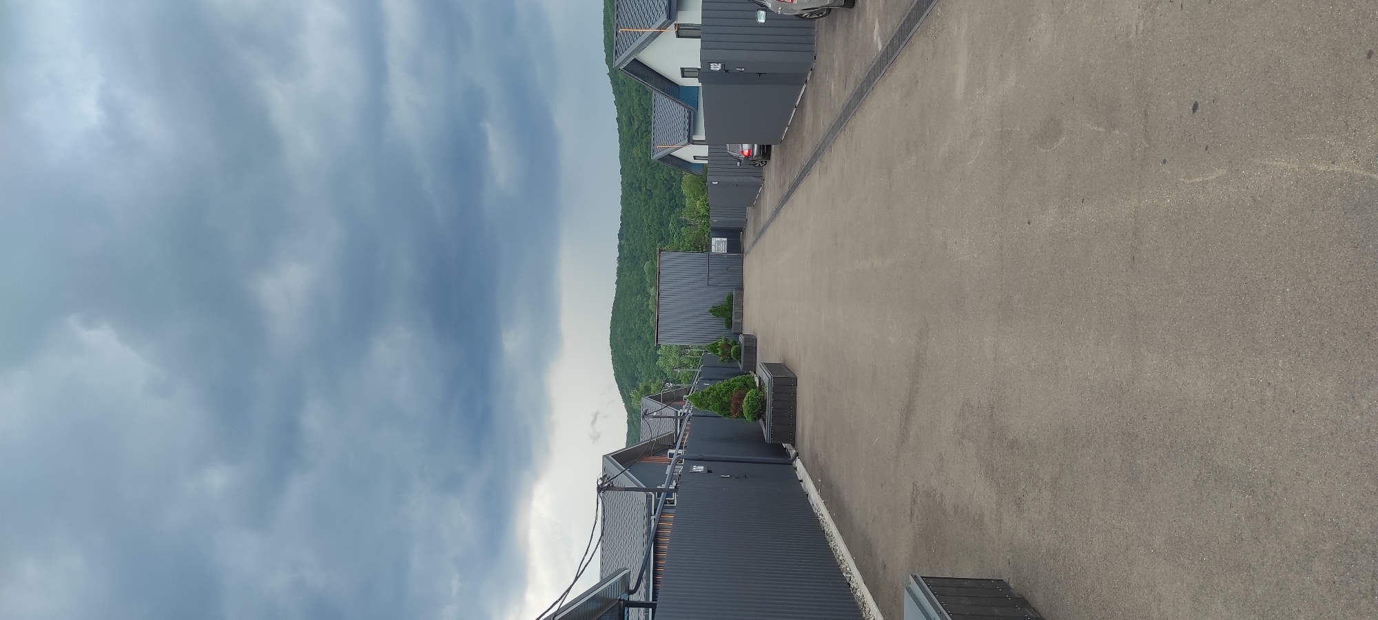 The image shows a row of modern, dark gray buildings with dark roofs, likely apartments or townhouses, situated on a hillside in Tulskiy, Russia. A paved driveway runs alongside the buildings. The buildings are in a linear arrangement, with some featuring white accents. Green trees cover the hillside behind the buildings. The sky is overcast with heavy, dark gray clouds, suggesting an impending storm or recent rain. There are several planters with small evergreen trees placed along the driveway. One of the buildings has a car parked in front of it. The overall scene conveys a sense of modern, somewhat minimalist architecture set against a natural, hilly landscape. There is no visible text in the image. The time of day appears to be daytime, but the weather conditions make it feel gloomy.