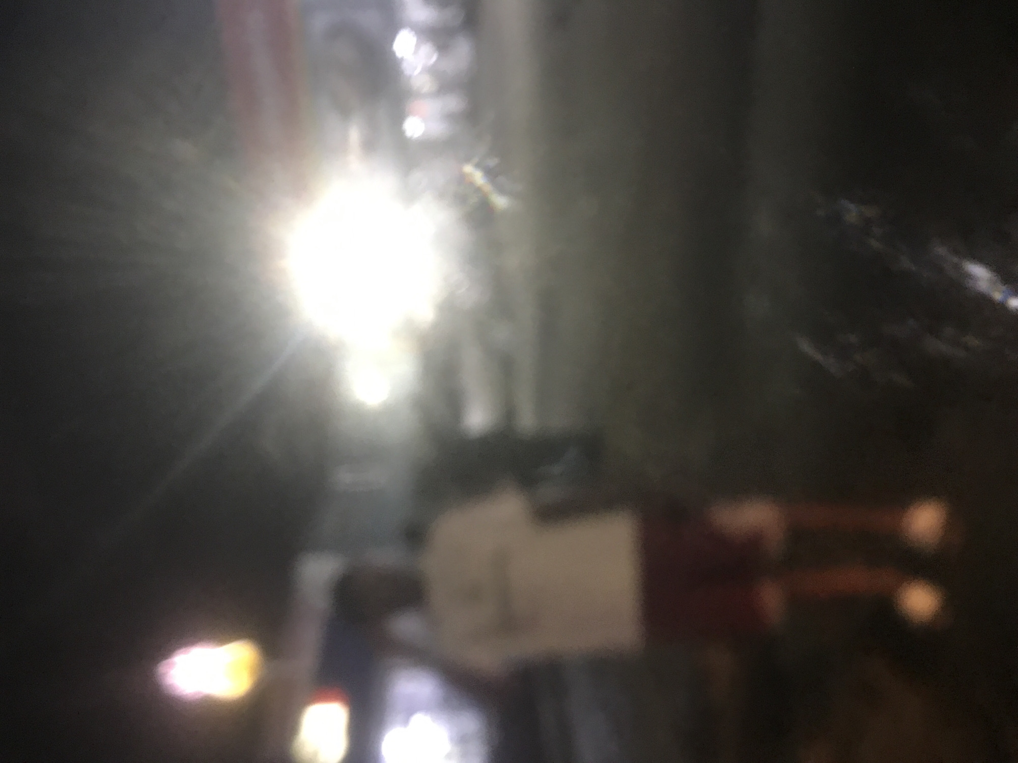 An outdoor, night time scene features a blurry figure of a person standing in the foreground. The figure is wearing a white t-shirt and dark red shorts, with white shoes. The person appears to be looking towards the left of the frame. Behind the figure, there are several out-of-focus lights, including a bright, diffused light source in the middle ground, which may be a street light or a vehicle's headlights. To the left, a vertical, elongated light source with pink and yellow hues is visible, resembling a lamp or a sign. In the background, other blurry shapes suggest the presence of buildings or structures, and possibly other people. The ground appears wet or reflective, indicating a potential for rain or recent moisture. The overall impression is of a dimly lit urban environment at night.