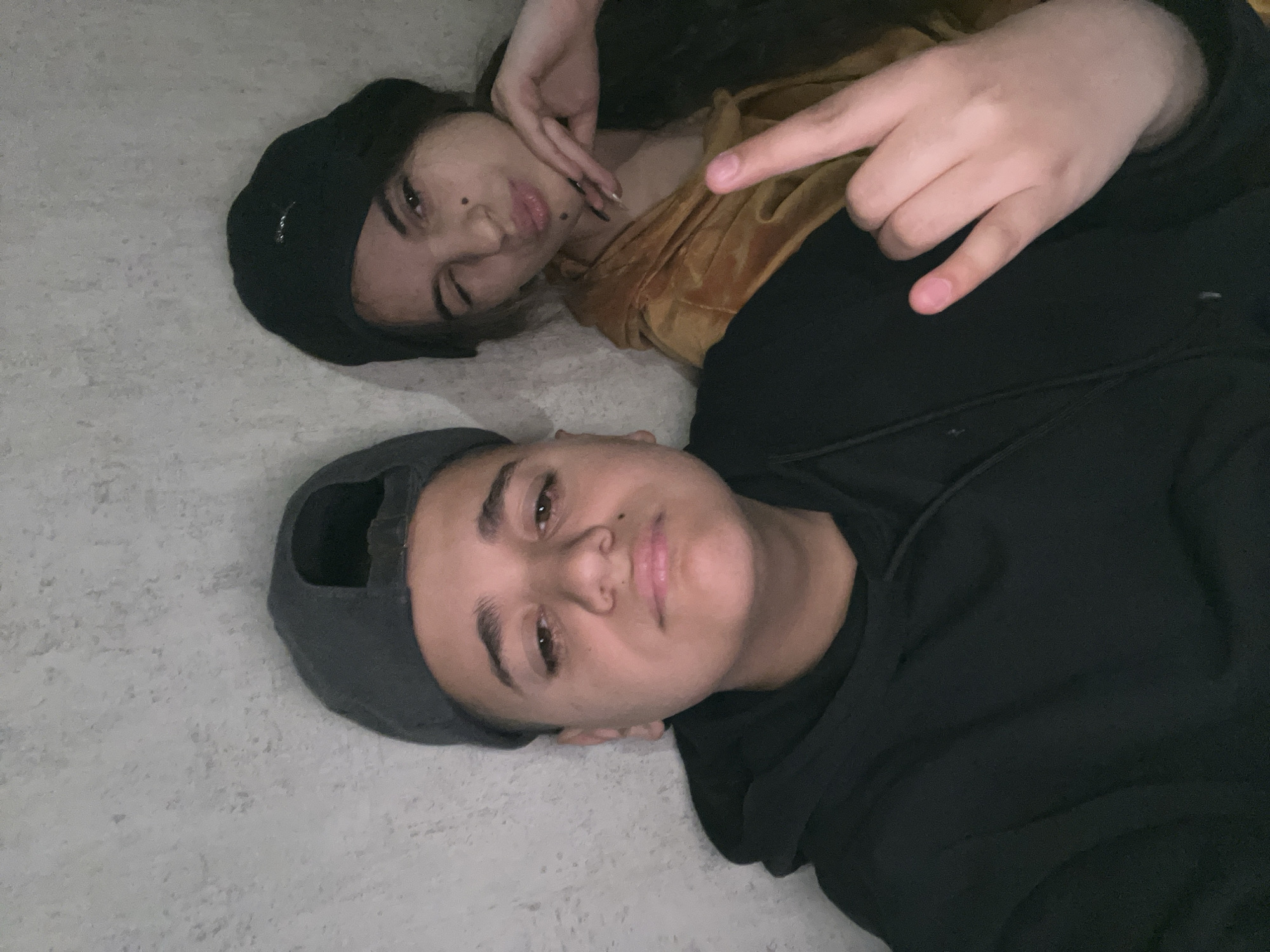 Two individuals are lying down, possibly on a floor, against a light-colored surface. The individual in the foreground wears a dark baseball cap and a black hooded sweatshirt. The second individual behind them is wearing a dark beanie, and a yellow garment. The person also has their fingers pointing at their throat and in the other hand making a horn gesture with their fingers. No evidence indicates a connection to the city of Lozarevo, Bulgaria.