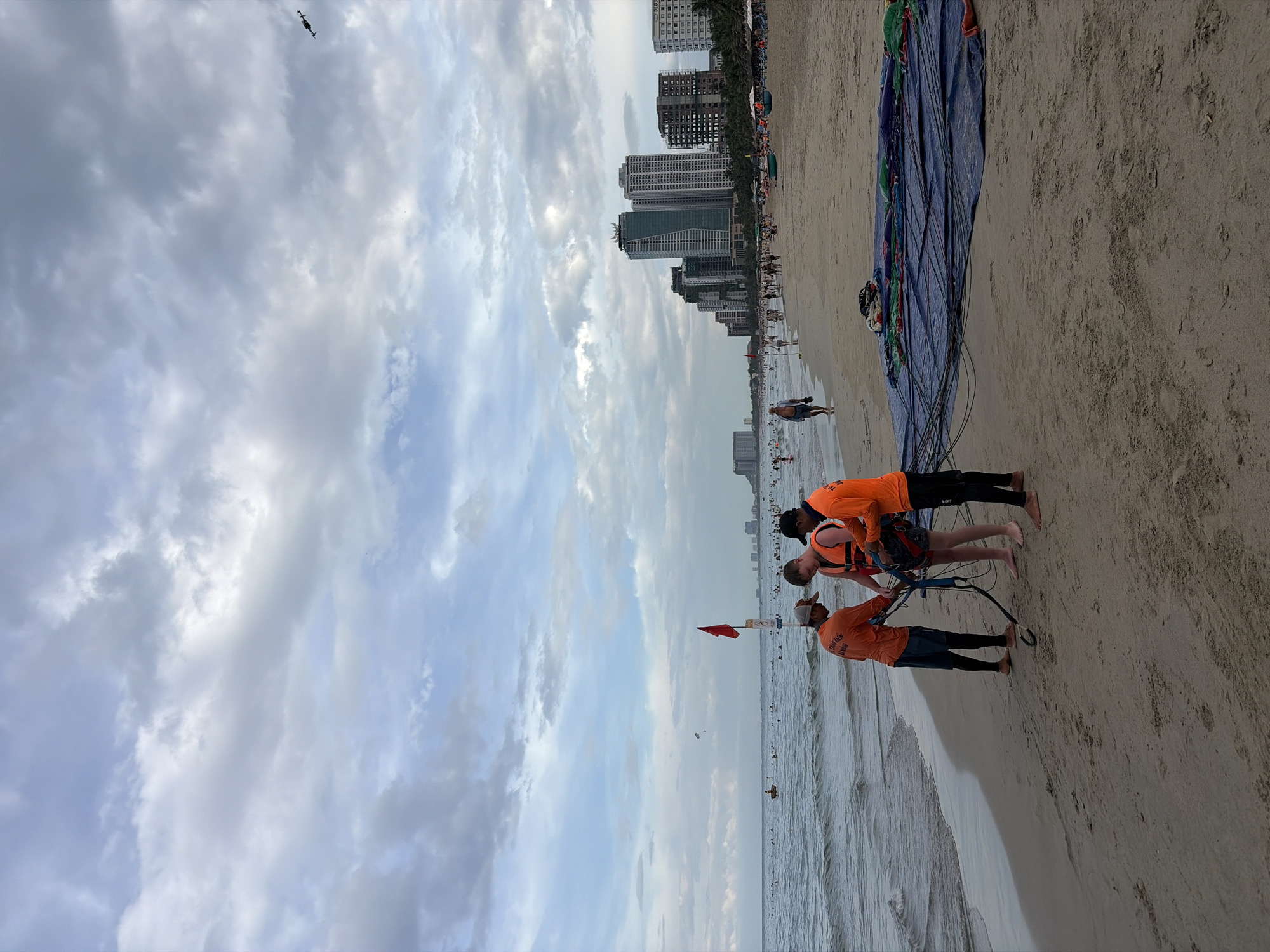 A beach scene in Da Nang, Vietnam features a group of people preparing for a watersport activity. In the foreground, three individuals are gathered on the sand near the water's edge. Two of them appear to be instructors or staff, wearing orange long-sleeved shirts and shorts, assisting a third person who is wearing a life vest and shorts. They are handling what looks like equipment for parasailing, with a large, colorful parachute spread out on the sand behind them. The ocean is calm with gentle waves.
In the background, the coastline is dotted with numerous beachgoers enjoying the water and the sand. A few small boats are visible in the sea. Further inland, a row of modern high-rise buildings stretches along the coast, characteristic of the Da Nang cityscape. The sky is overcast with dramatic clouds, suggesting a potentially changeable weather day. The overall atmosphere appears to be one of recreation and a bustling tourist destination.