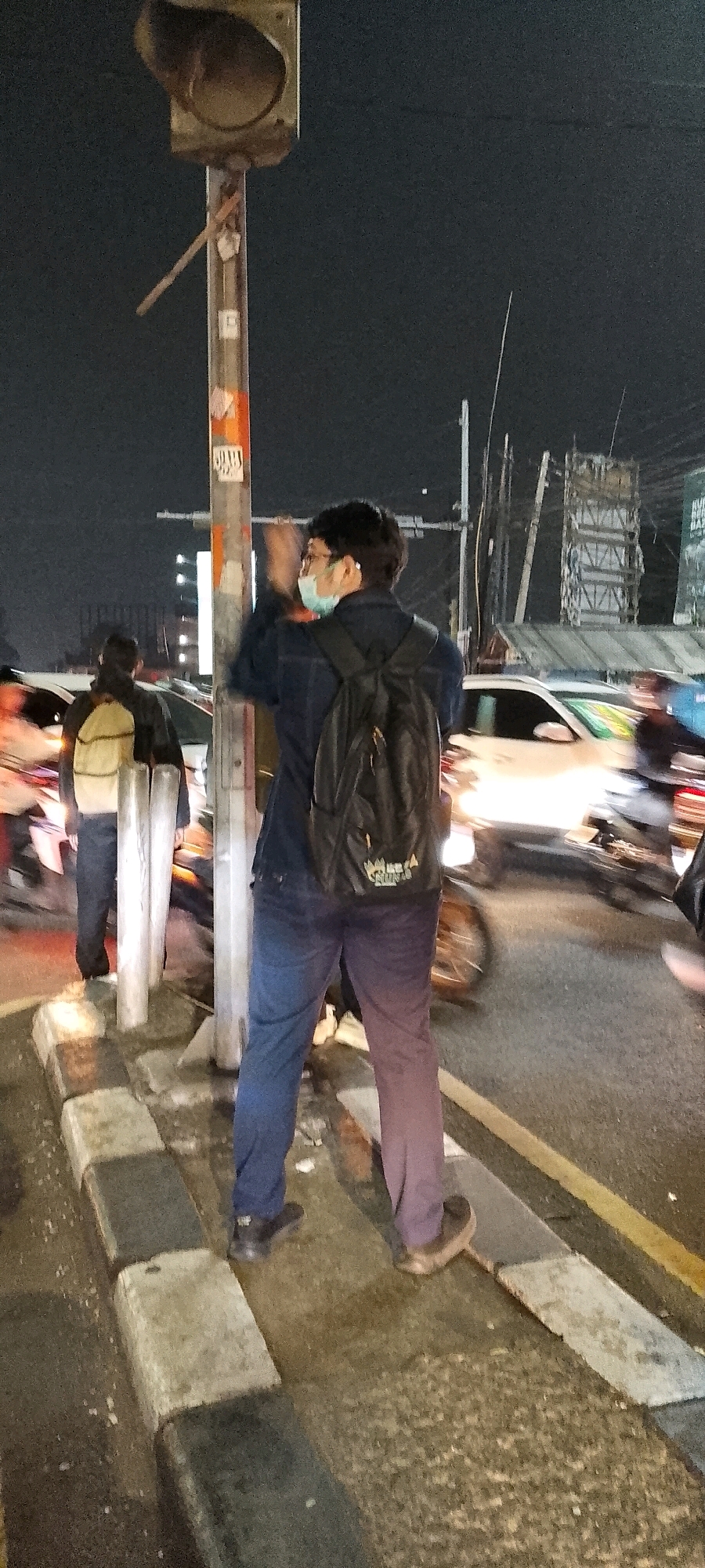 The image captures a night scene on a street, likely in Depok, Indonesia, as indicated by the context. A traffic light pole stands prominently in the foreground, adorned with stickers. A man wearing a blue jacket, blue pants, a face mask, and a black backpack is standing beside the pole, with his right arm raised and seemingly in motion, possibly interacting with someone or something out of frame. His left hand is not visible. Behind him, another person with a light-colored backpack is walking away from the camera, also near the traffic light pole. The street is wet, suggesting recent rain, and several vehicles, including motorcycles and cars, are visible in motion, indicated by light trails and motion blur. The background shows streetlights and buildings, typical of an urban environment at night. The overall atmosphere is that of a busy intersection.