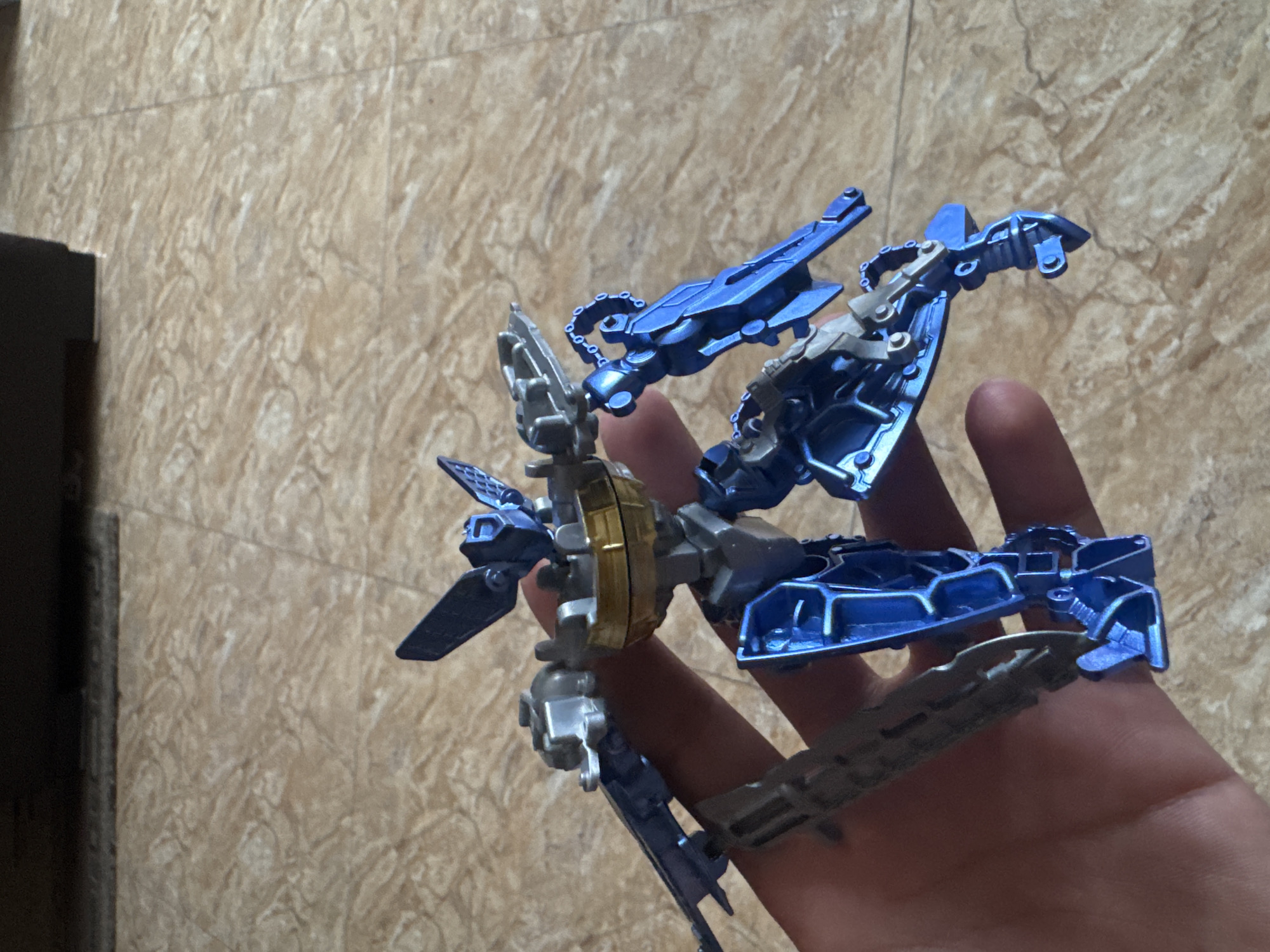A hand holds a disassembled toy robot, primarily blue and silver in color. The background is a light brown wall with a patterned texture, possibly a tile wall. There is a dark object visible on the left edge of the frame. The image was not taken in Yerevan, Armenia, as that information cannot be deduced from visual elements.