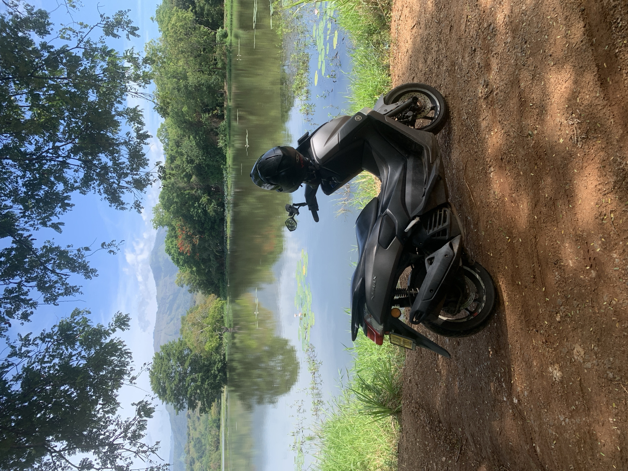 In Buduruwagala, Sri Lanka, a grey scooter is parked on a dirt path next to a body of water. The scooter is positioned in the foreground, angled slightly towards the left. A black helmet is placed on the handlebars. The water's surface reflects the surrounding lush greenery and distant hills under a clear blue sky with scattered clouds. Lily pads and aquatic plants are visible on the water. The environment is natural and serene, with trees and foliage lining the banks of the water.