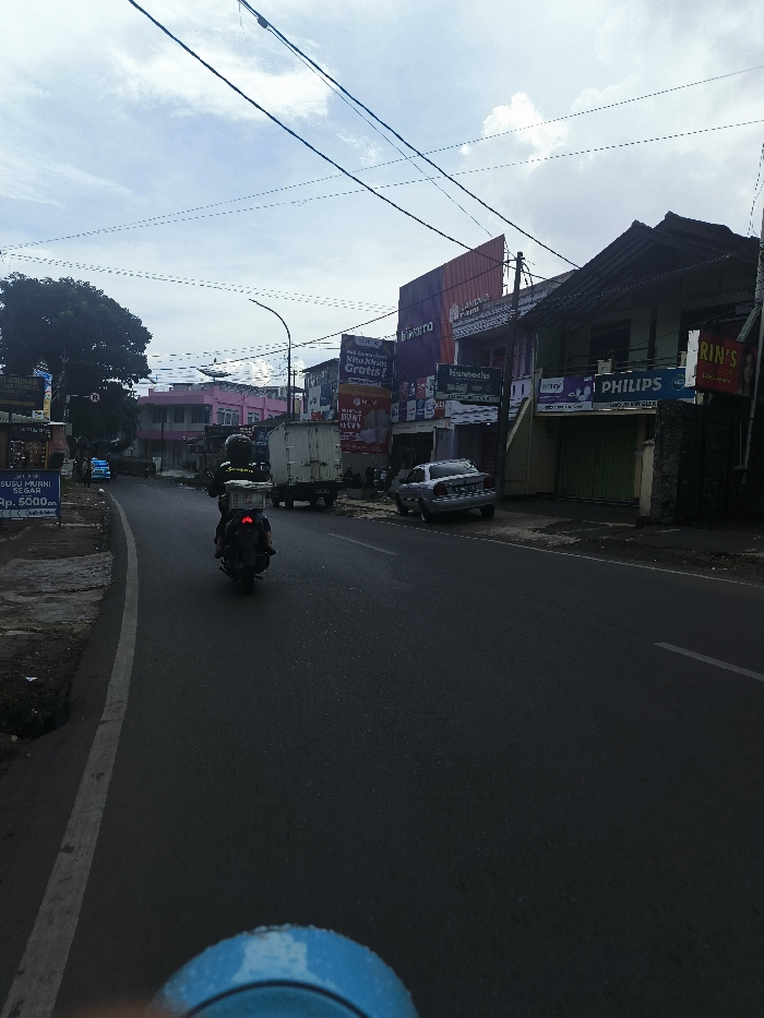 The image shows a street in Lembang, Indonesia. The coordinates, -6.816 latitude and 107.619 longitude, indicate a location in a residential area. The scene is dominated by a two-lane road with a white dividing line. A motorcycle with a rider is visible, moving away from the camera on the right lane. The street is lined with shops on the right side, with signs for businesses like "Kivero," "Hanroaches," "Philips," and "Rin's Laundry." The buildings have a light gray color and appear to be made of brick or concrete. There are several power lines stretching across the sky above the street. On the left side of the road, there are more buildings, a few cars, and a small sign for a store that sells vegetables. The overall scene is one of typical Indonesian suburban life. The sky is a light blue with some clouds. The weather appears to be overcast.
