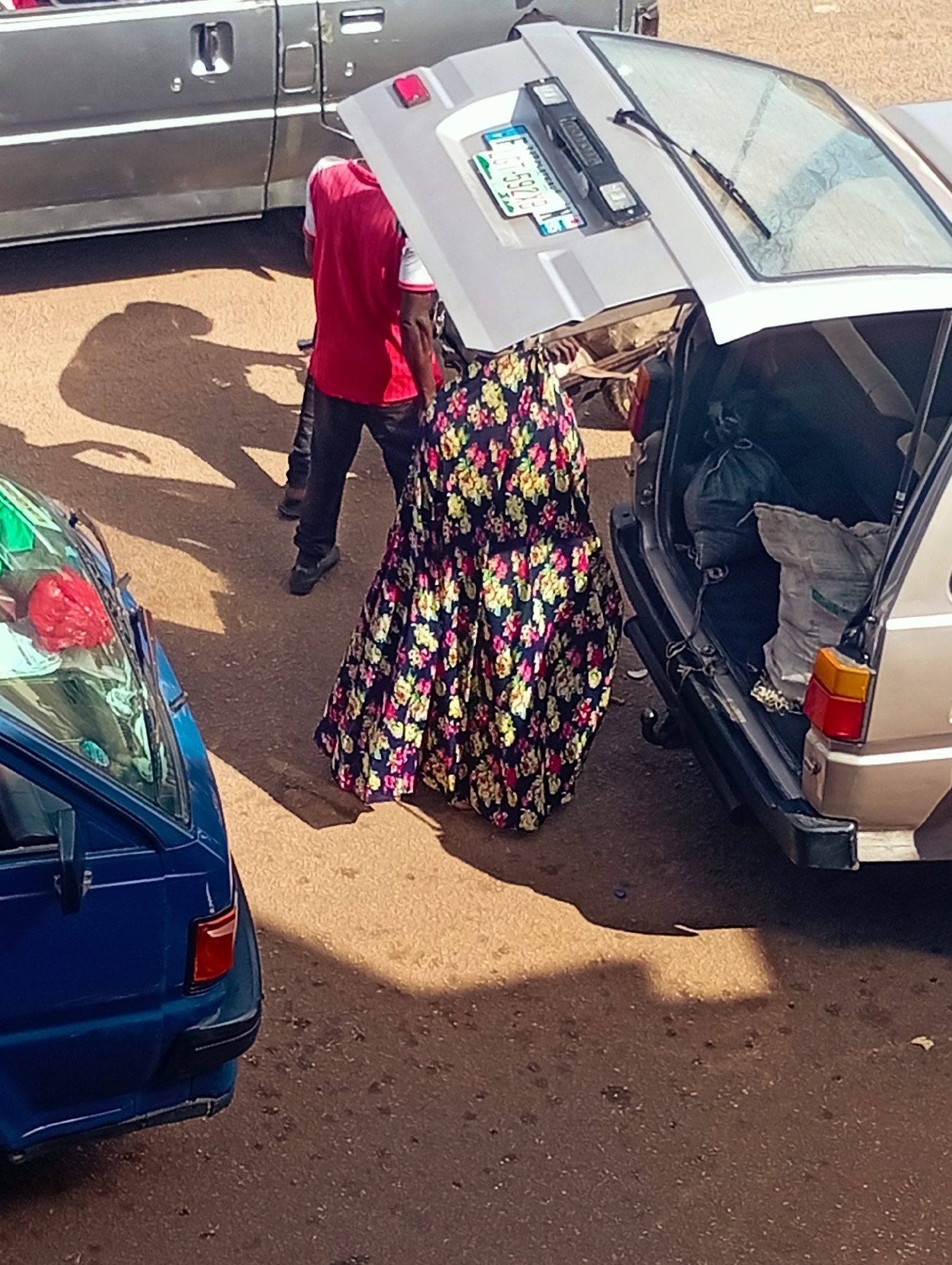 In Bukuru, Nigeria, a busy street scene unfolds with vehicles and people. The foreground features a blue vehicle, its driver's side mirror visible. Adjacent to it, a woman is walking away from the viewer, her long, floral-patterned skirt creating a voluminous silhouette. Her attire suggests cultural significance or personal style. Behind her, a car with its trunk open reveals a jumble of items, including bags and possibly luggage. A man in a red shirt and black trousers stands near the open trunk, appearing to be involved in the loading or unloading process. His posture and engagement suggest a purposeful activity. In the background, another vehicle, possibly a truck or van, is parked. The ground is paved with asphalt, worn by traffic. Sunlight casts long shadows, indicating a time of day when the sun is at a lower angle. The overall impression is one of everyday life in a Nigerian city, with a focus on commerce or transportation.