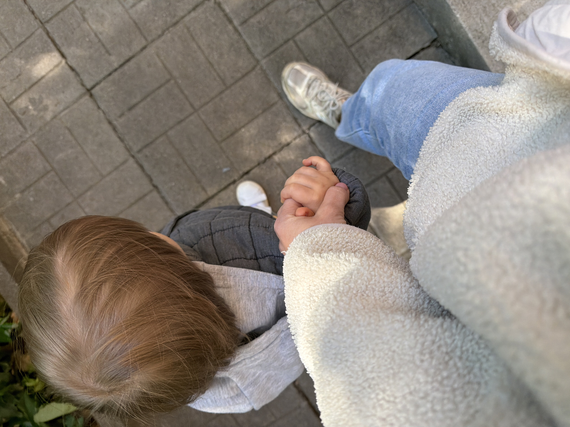 A high-angle shot shows an adult and a child holding hands on a paved surface. The child is to the left, with visible light brown hair and wearing a gray quilted jacket over a light gray sweater. The adult is to the right, wearing blue jeans, a white sneaker, and a white fuzzy jacket. The paved surface appears to be made of gray rectangular blocks. It is difficult to determine the location within Sofia, Bulgaria without additional visual cues.
