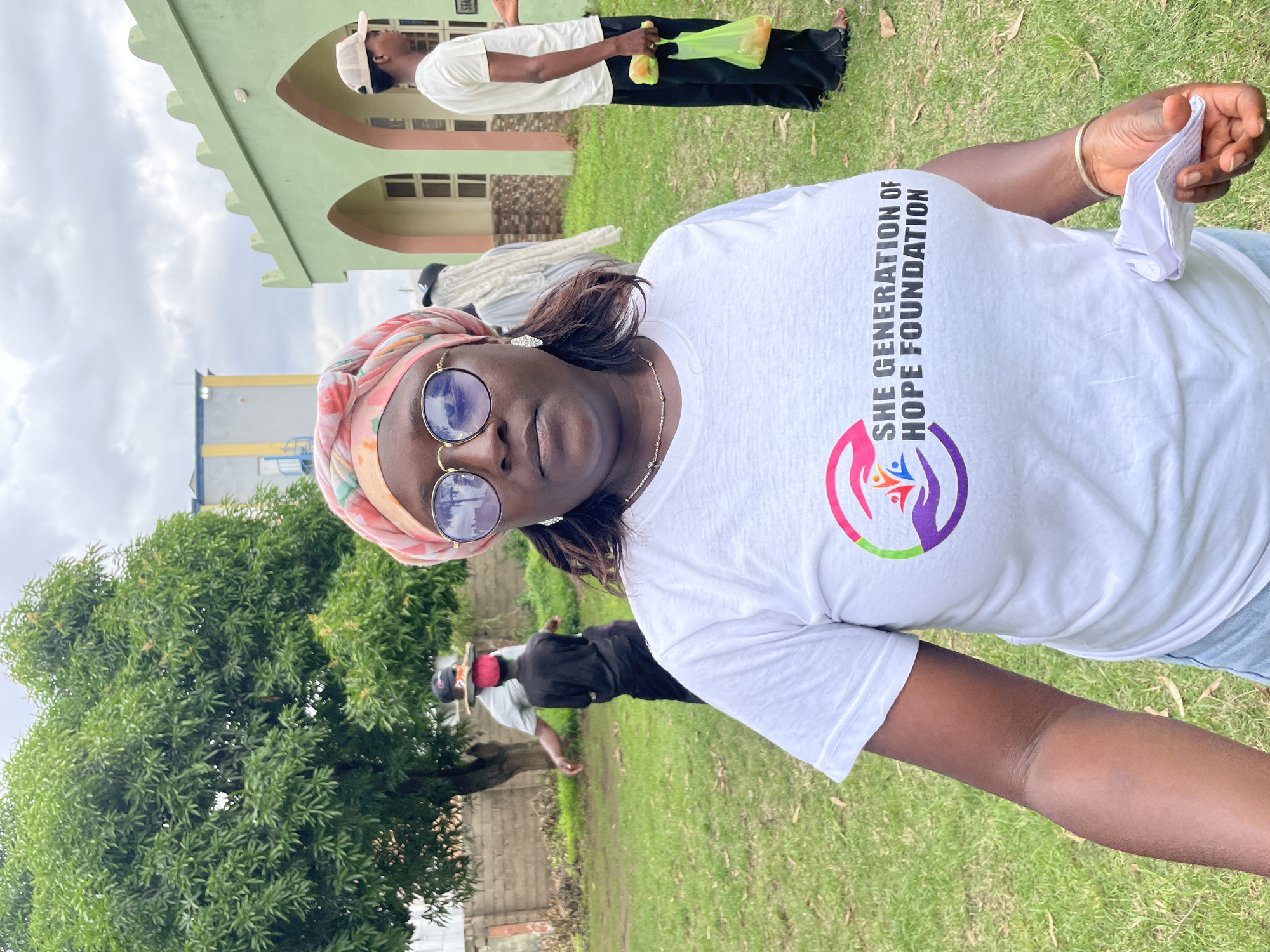 A group of people are gathered outdoors on a cloudy day in Unguwar Shanu, Nigeria. The main subject is a woman in the foreground, wearing a white t-shirt with the text "SHE GENERATION OF HOPE FOUNDATION" and a logo. She has a headscarf on and is wearing sunglasses. She is holding a crumpled piece of paper in her right hand.
In the background, a person wearing a white t-shirt and black pants is holding a bright yellow bag. Further in the background, a building with a light green facade and arched windows is visible, suggesting a public or institutional setting. There are also trees and grassy areas, indicating an outdoor environment. The overall mood appears casual, possibly related to a community event or outreach program organized by the foundation mentioned on the t-shirt.
