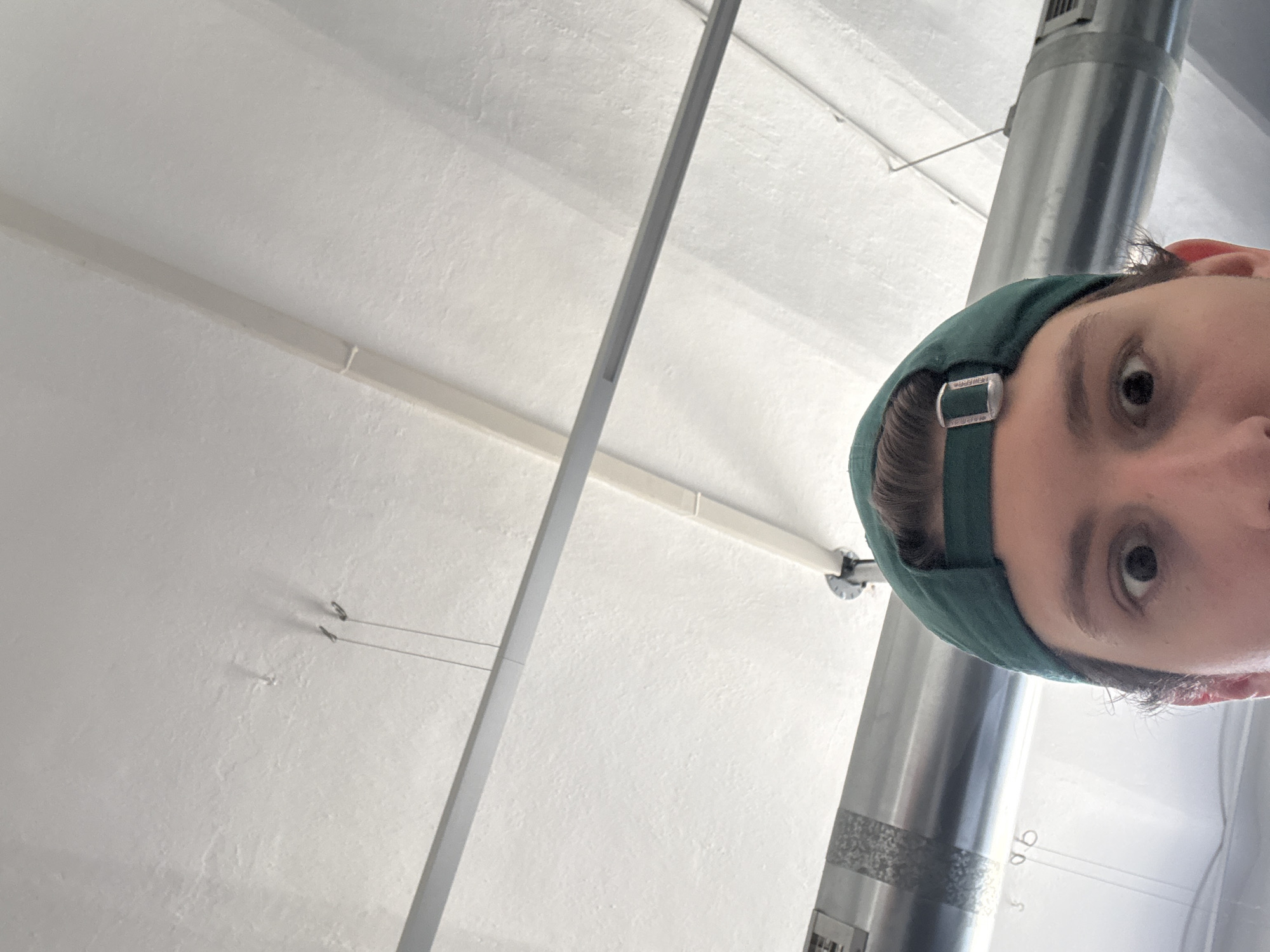 A first-person, upward-facing perspective of a person wearing a green baseball cap with a silver buckle on the back adjuster, with their face partially visible in the upper right portion of the frame. The person is looking directly at the camera.
The background consists of a white, industrial-style ceiling with fluorescent lighting fixtures, metal support beams, and a large, cylindrical metal duct running vertically through the frame. The location appears to be indoors, possibly a factory, warehouse, or commercial space in an industrial area. There is no specific landmark visible to place this image in Milan, Italy.