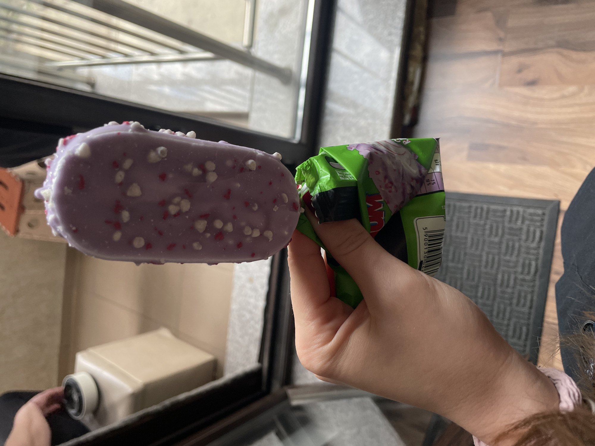 A person is holding a purple popsicle with white and red sprinkles in one hand and the wrapper of the popsicle in the other hand. The popsicle is rectangular with rounded edges. The wrapper is green and purple with some black tape. The background includes a window with a metal grate, a wooden floor, and a gray mat. A white box is visible through the window. The location appears to be indoors, but the specific location in Sofia, Bulgaria cannot be determined from the visual information provided.