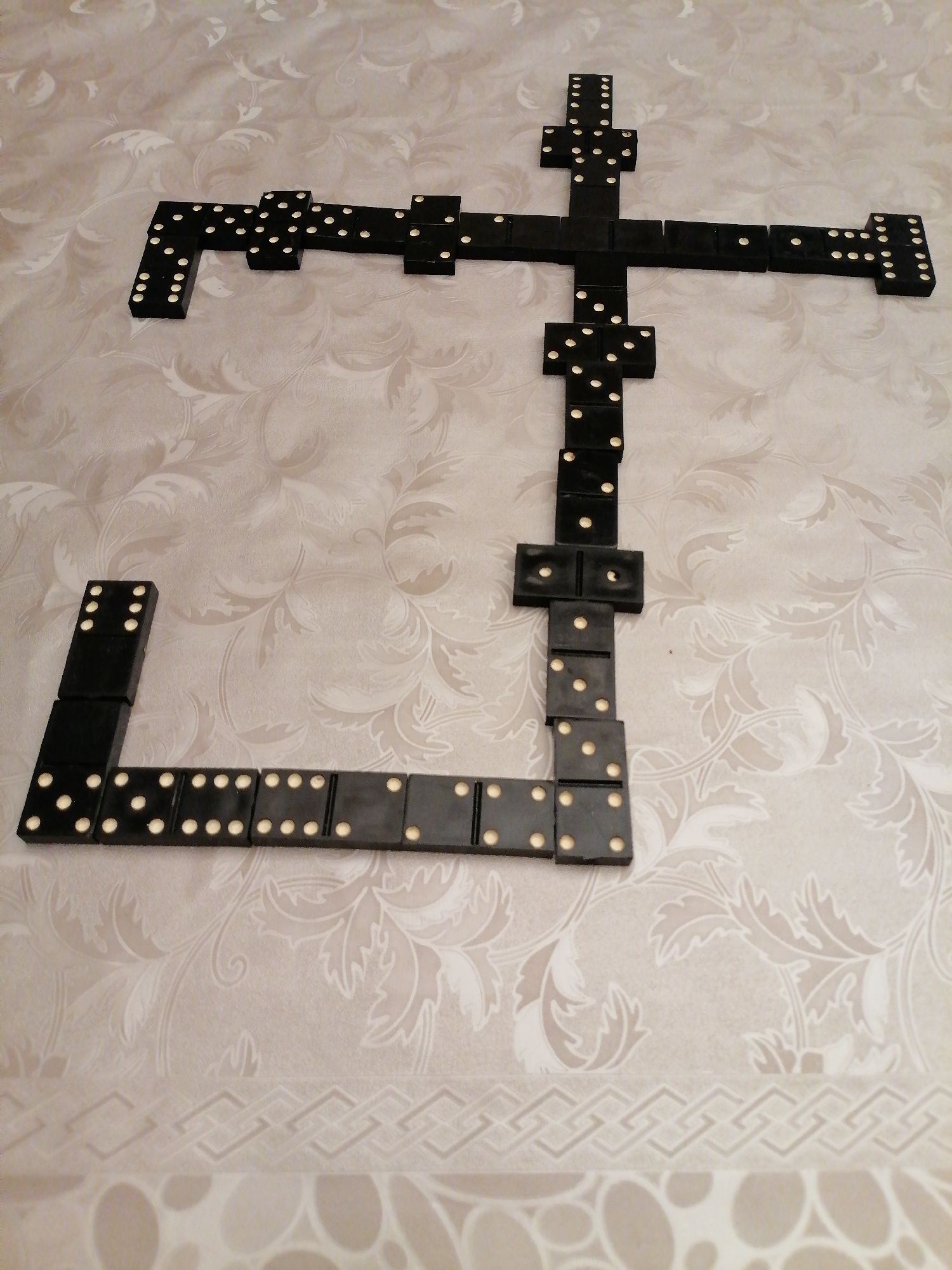 The image shows a close-up, eye-level, overhead shot of a domino game in progress on a textured, light-colored tablecloth with a floral pattern. The dominoes are black with white dots and are arranged in a cross-like formation. The arrangement appears to be in the process of a game, with multiple dominoes forming the body of the cross and extending outwards. The lighting is soft and diffused, suggesting an indoor setting. There are no people visible in the image, and no contextual clues suggest a specific location within Rustavi, Georgia, or any other city. The focus is entirely on the dominoes and the immediate surface they are on.