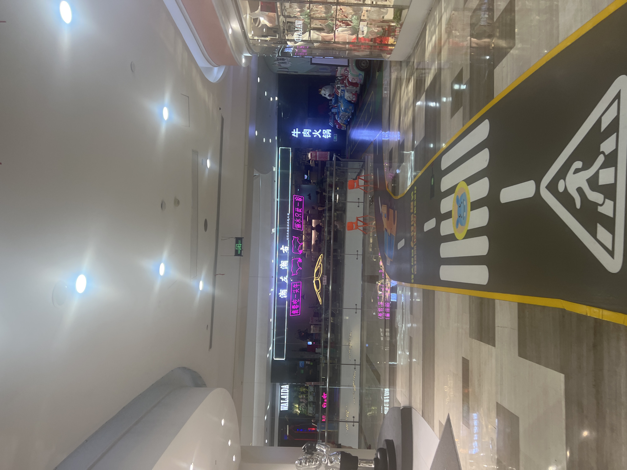 The image shows the interior of a shopping mall in Liandu District, China. In the foreground, a children's play area is designed to resemble a road with a crosswalk. The road features white painted lines for lanes and a pedestrian crossing symbol with a cartoon animal. In the middle ground, there is a row of colorful children's ride-on cars. Beyond the play area, a glass partition separates the walkway from a dining establishment with neon signage in Chinese characters, including "牛肉火锅" (beef hot pot). To the left, a store with the "WALAIDA" logo is visible. The mall environment is illuminated by overhead lights and reflects off the polished floor. In the background, more shops and dining areas can be seen.