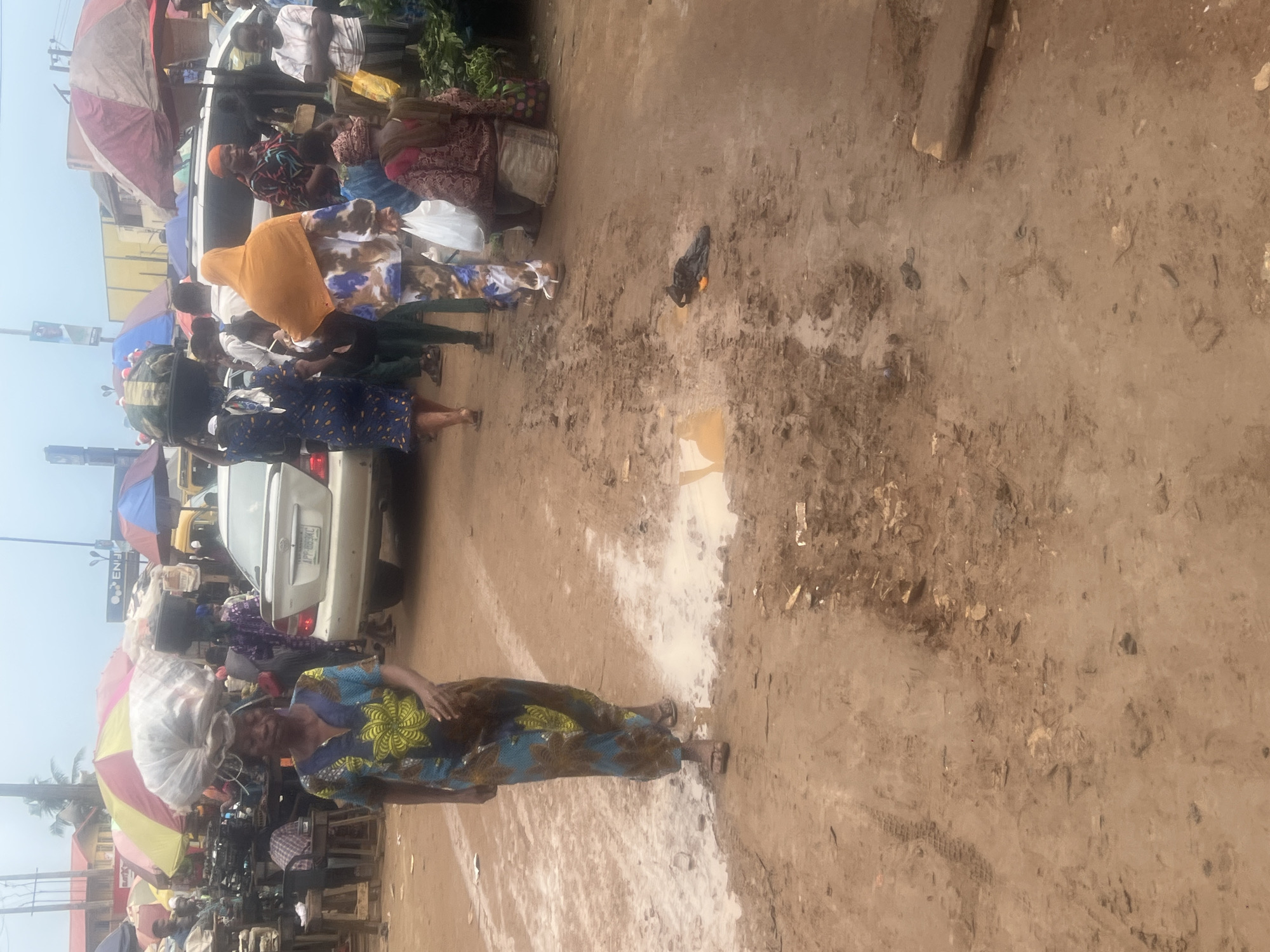 The image depicts a bustling street scene in Egan, Nigeria. A woman in a patterned blue and yellow dress carries a large white load on her head, walking on a muddy, uneven ground. A white car is parked nearby, with people gathered around it. Several individuals carry items on their heads, and colorful umbrellas provide shade from the sun. The environment appears to be a marketplace, with various vendors and pedestrian traffic. The overall scene conveys a sense of daily life and commerce in a crowded urban environment.