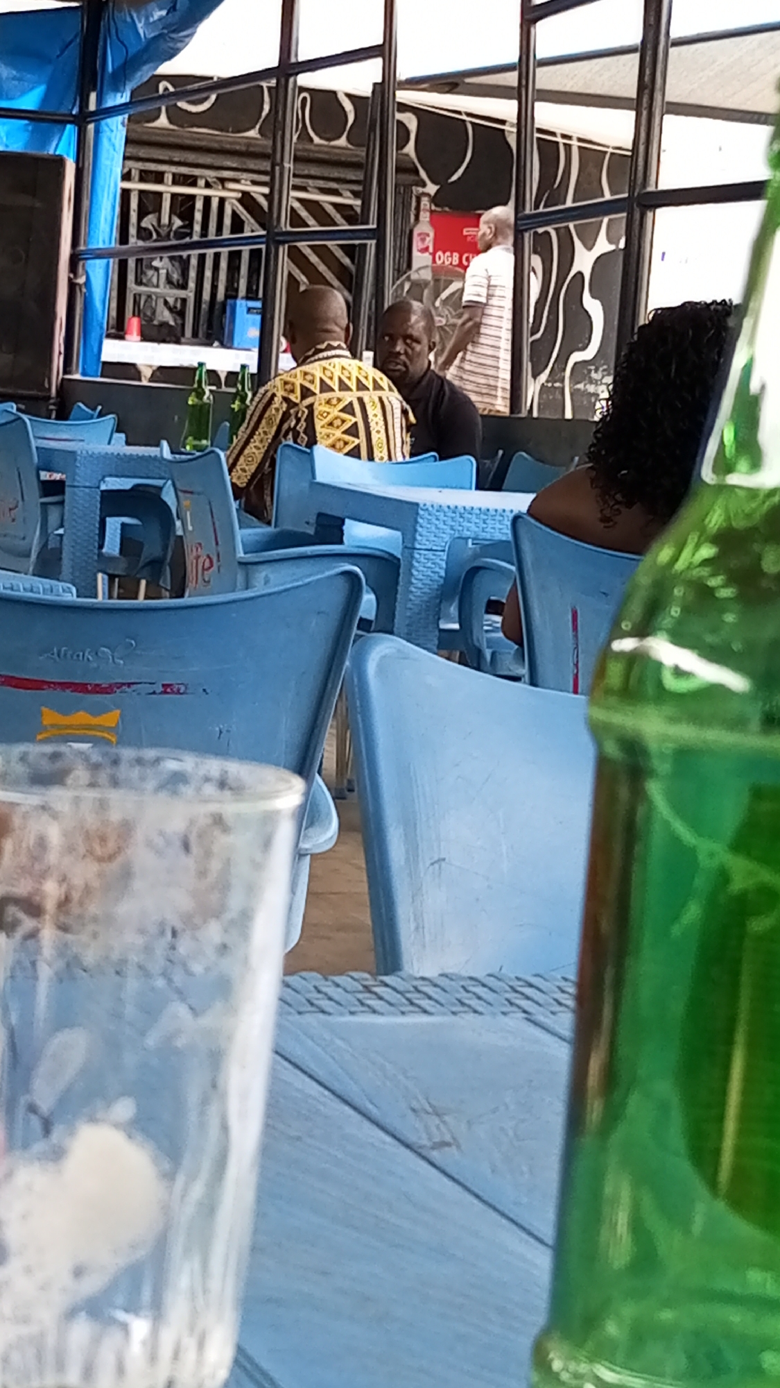 The image shows an outdoor scene, likely a restaurant or bar in Sabon Tasha, Nigeria. There are blue plastic chairs and tables. Several people are seated, including two men facing away from the camera, one wearing a patterned shirt. A woman with curly black hair is seated at another table. In the background, a man in a striped shirt stands near a cooler. A glass with some residue is in the foreground on the left. A green beer bottle is prominent on the right. The backdrop includes a building with a black and white decorative facade.