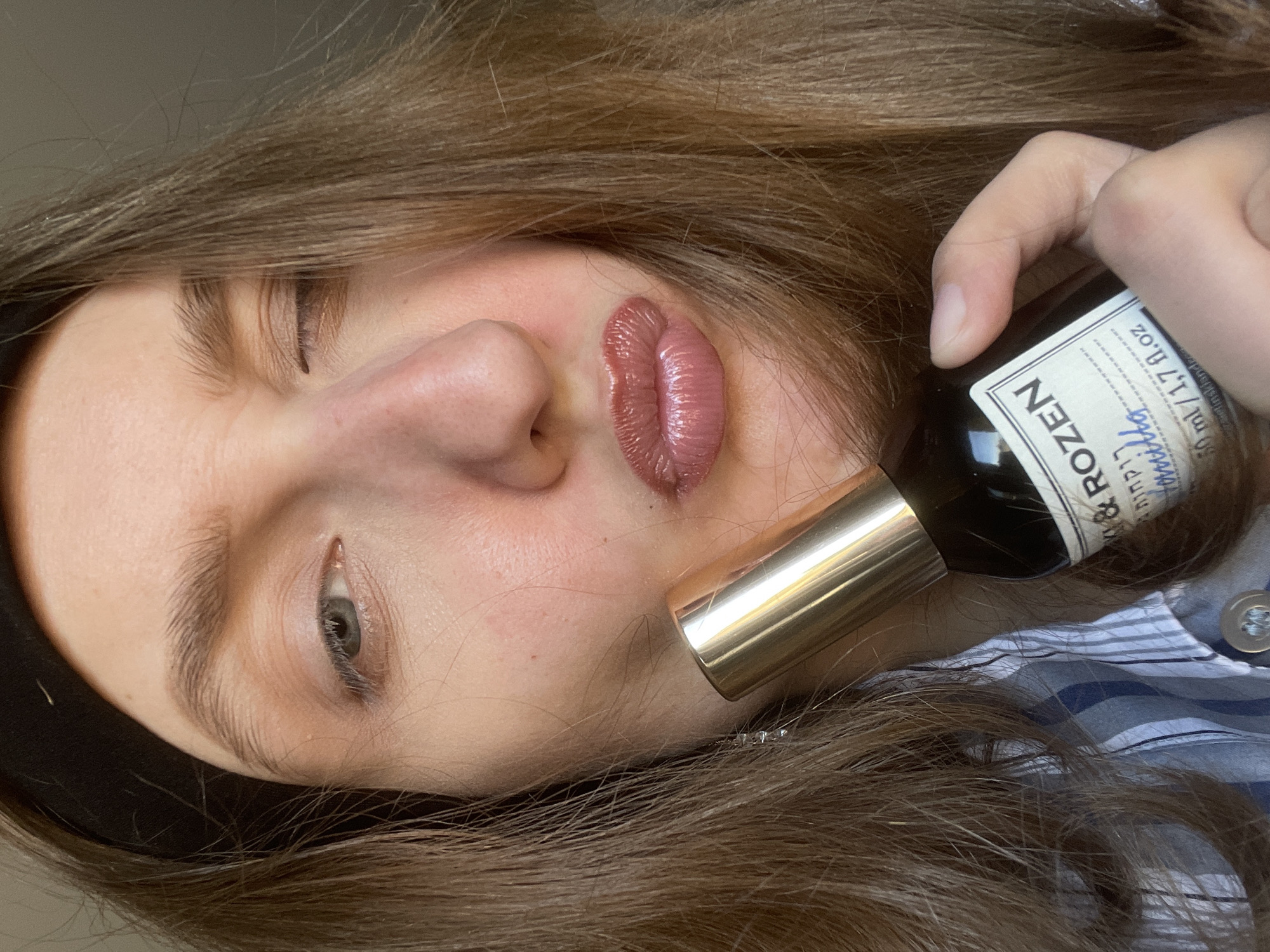 A light-skinned person with brown hair and closed eyes holds a bottle labeled "ROZEN" in their right hand. The bottle is dark with a white label. A gold-colored cylindrical object is positioned near the person's cheek. They wear a dark headband, and a shirt with a blue and white pattern is visible. The image is rotated sideways. There is no indication that the image was taken in Podshivalovo, Russia.