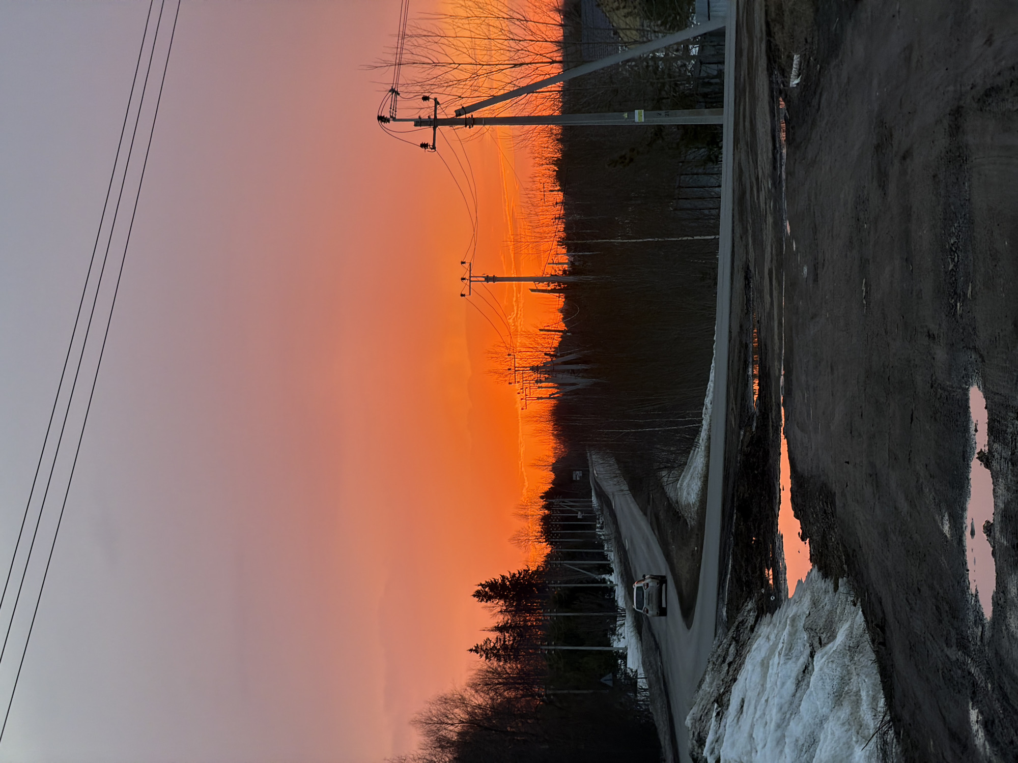 The image depicts a road in the vicinity of Izhevsk, Russia, during a sunset. The sky is a gradient of color, ranging from gray to orange. Power lines and poles are visible along the road. Trees line the roadside. A vehicle is traveling on the road. Melting snow is present along the side of the road, and a puddle is on the unpaved section.