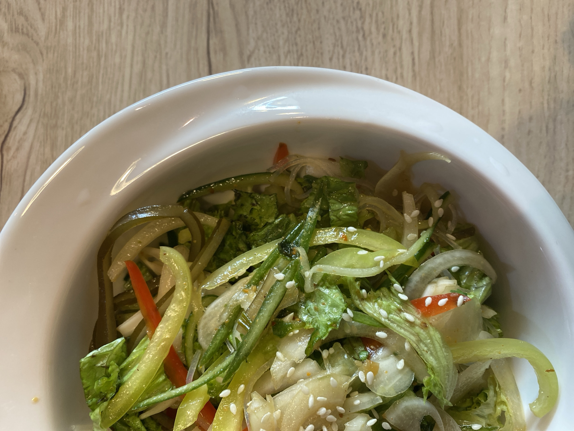 A close-up, overhead shot shows a white bowl filled with a colorful salad. The salad contains thinly sliced green and red peppers, cucumber, lettuce, onion rings, and what appears to be glass noodles or kelp. White sesame seeds are sprinkled over the top. The bowl is placed on a light-colored wooden surface, possibly a table. The background is slightly blurred, with the wooden texture continuing. No people are visible in the image. The scene is indoors, likely in a restaurant or dining setting.