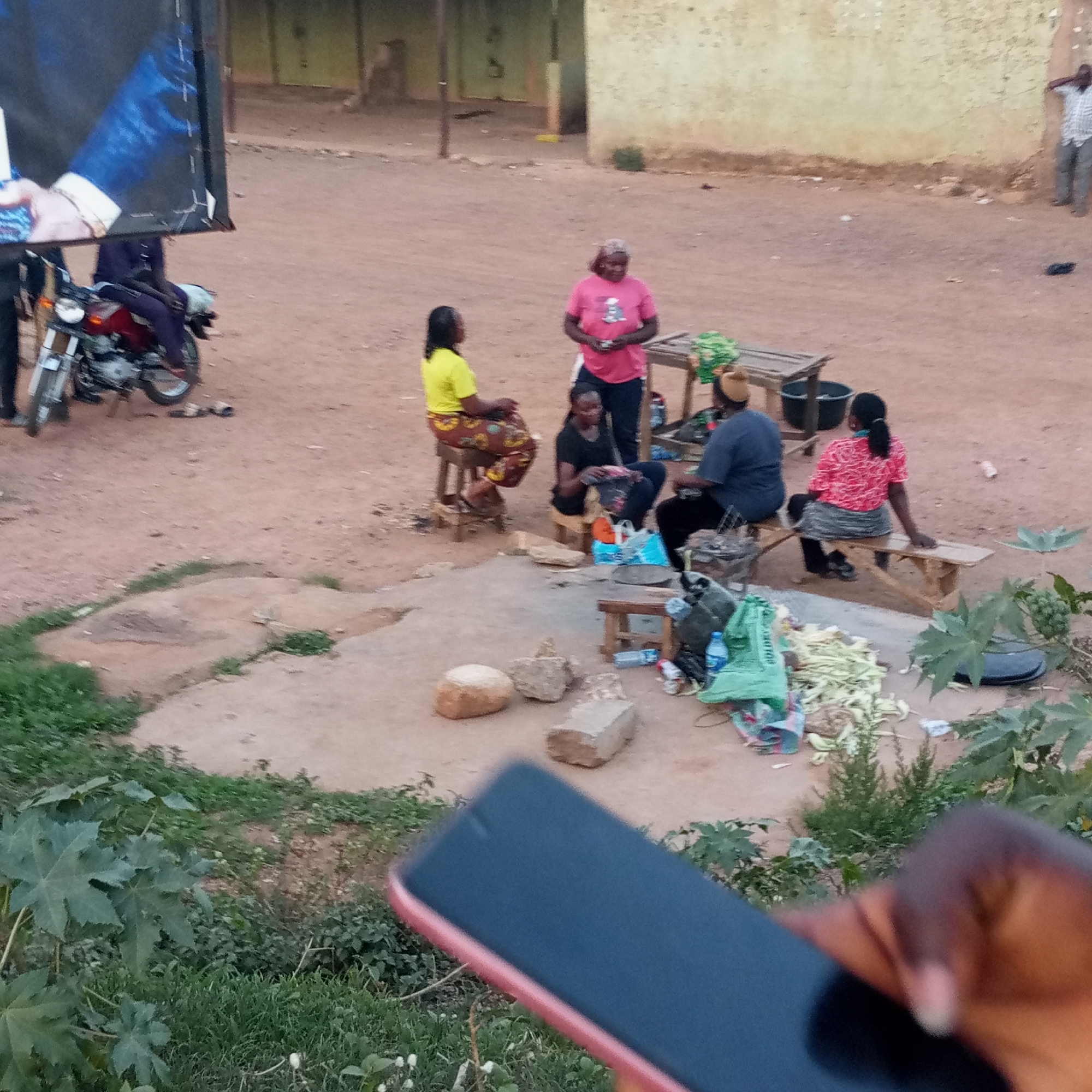 The scene is set in Kuru Dungus, Nigeria. Five women are gathered in a dirt clearing, likely engaged in commerce or social interaction. One woman wears a yellow shirt and patterned skirt while sitting on a small stool. Another woman is standing, wearing a pink shirt. A third woman sits in a black shirt and dark pants. A fourth woman in a grey shirt and hat is seated on a stool. The fifth woman, wearing a red patterned shirt and dark skirt, sits on a wooden bench. Various items including what appear to be vegetables or foodstuffs, along with bags and containers, are situated on the ground around the women. There are also several rocks and small wooden stools. In the background, there is a man standing against a light-colored wall. A motorbike is parked to the left of the women, adjacent to a billboard featuring a man in a suit. In the foreground, a hand is holding a dark-colored smartphone with a pink or red border. The surrounding vegetation consists of green plants and grass. The ground is primarily dirt or packed soil.