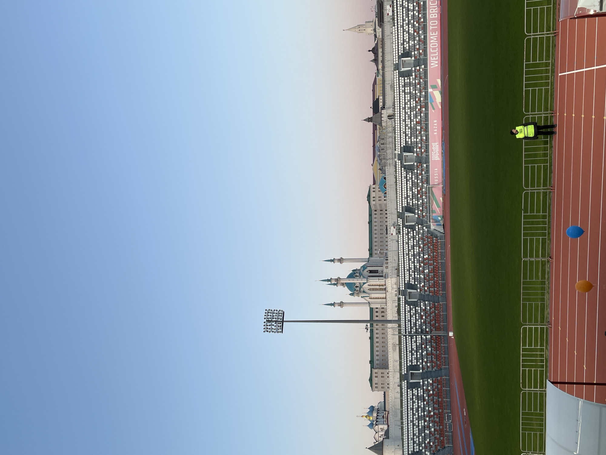 The image captures Kazan, Russia, with a stadium and city landmarks under a clear sky. The stadium features green grass and a running track, with a person in a yellow safety vest visible. Nearby are architectural structures, including the Qolşärif Mosque, which is recognizable by its turquoise domes and minarets. Adjacent to the stadium, stands of seating are visible. In the background, various city buildings can be seen.