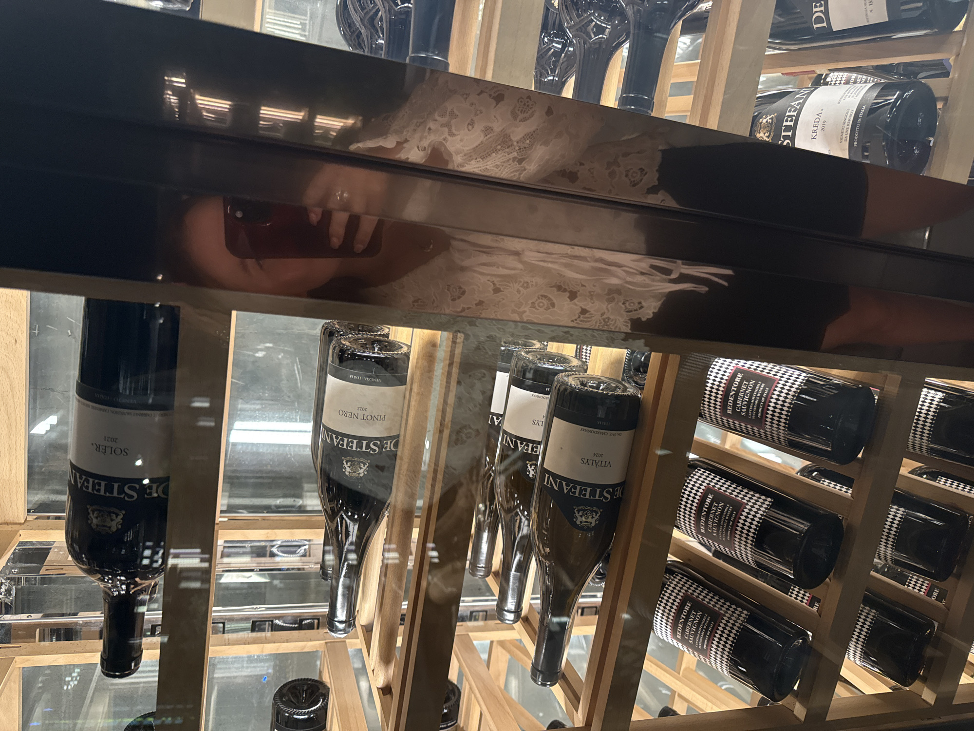 A woman, partially visible in reflection, holds a red smartphone in her right hand, using it to take a selfie. She is wearing a white lace top, with her left hand resting on a dark, reflective surface. The reflection shows her face and upper body. The woman is positioned in front of a wine rack filled with numerous bottles of wine. The wine rack is made of light-colored wood and has multiple shelves. The bottles are of various brands and types, including red and white wines. Some labels are visible, such as "DE STEFANI" and "REDENTORE CABERNET SAUVIGNON." The overall environment appears to be an indoor retail space, possibly a wine shop or a restaurant with a wine display. The image's composition suggests the woman is inside a refrigerated wine cabinet or a similar display unit with glass doors, as indicated by the reflections and the organized rows of bottles. The specific location in Yiwu, China, is not explicitly indicated by any visual cues in the image itself.
