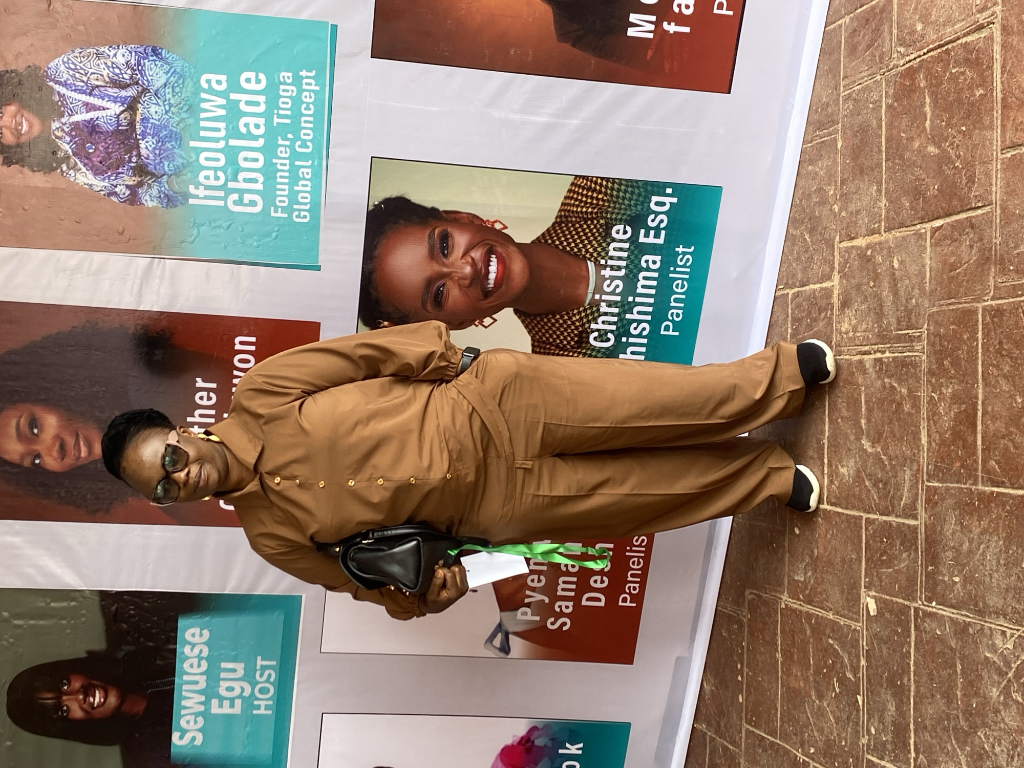 A medium shot captures a woman standing on a tiled floor in front of a banner displaying speaker profiles. The woman, appearing to be in her 40s or 50s, is wearing a brown button-down shirt and matching wide-leg trousers. She has on large, dark-rimmed glasses and gold hoop earrings. A black handbag is slung over her left shoulder. A white tag with a green ribbon hangs from the handbag strap.
The banner behind her features several printed portraits of women with their names and titles. From left to right, the visible text includes "Sewuese Egu HOST," and a partially obscured name followed by "won." To the right of that is "Ifeoluwa Gbolade Founder, Tioga Global Concept." Further to the right is "Christine Nishima Esq. Panelist." Another section of the banner shows "M" followed by "fa" and "P."
The tiled floor is reddish-brown and appears worn, with visible grout lines and some dust or debris. The lighting suggests an outdoor or well-lit indoor setting. The overall impression is of a professional event, possibly a conference or seminar, taking place in Jos, Nigeria, given the context of the banner's content and the general appearance of the environment.