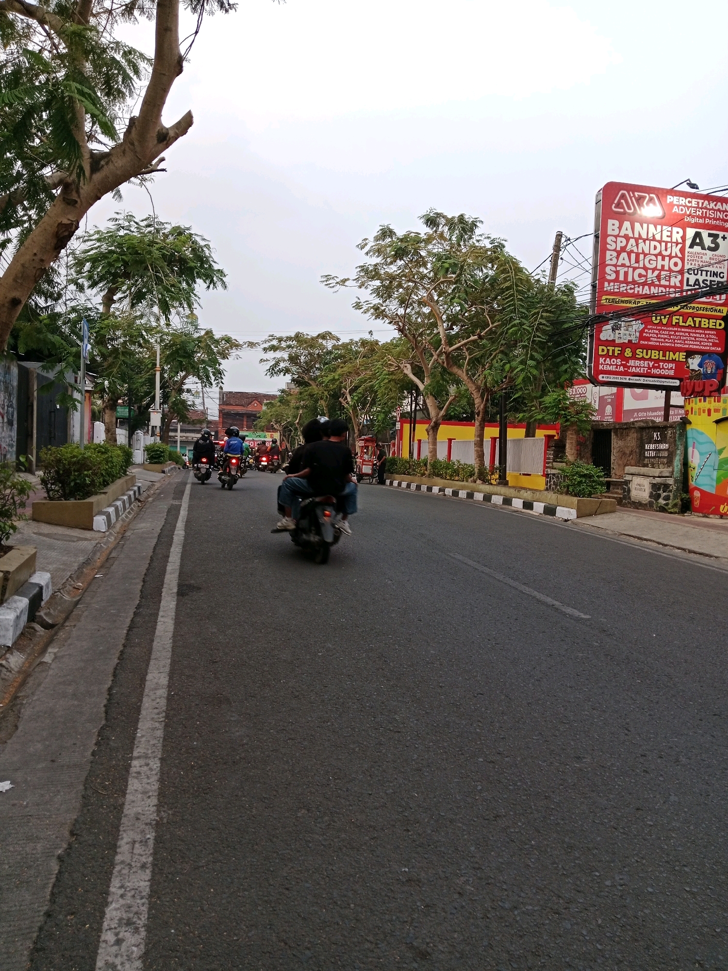 The image depicts a street scene in Cianjur, Indonesia, characterized by overcast skies and a gray asphalt road. On the right side of the road, a large red and white billboard advertises "PERCETAKAN ADVERTISING" services, featuring offerings like banners, stickers, and merchandise printing, along with contact information. Adjacent to the billboard, the building displays vibrant graffiti art. A row of trees lines the left side of the street, with a sidewalk featuring a distinctive black and white striped curb. Several people are riding motorcycles on the road; one motorcycle in the foreground carries two individuals, with the passenger seen from the back. Other motorcycles are visible further down the street, forming a small procession. The environment appears to be urban, with buildings and lush greenery contributing to the streetscape.