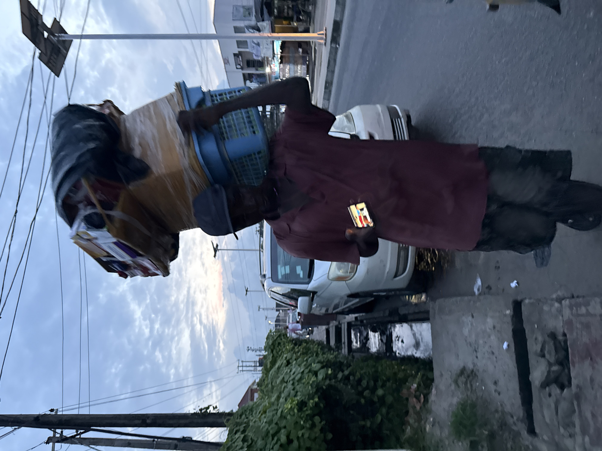 In Ado, Nigeria, a man carrying a large bundle of goods on his head walks along a street at dusk. The bundle, wrapped in plastic, rests on a blue plastic basket. He wears a maroon short-sleeved shirt, dark pants, and a black cap. In his right hand, he holds a smartphone displaying a colorful image. A white van is parked on the right side of the street, and a concrete drain runs alongside the road, lined with lush green bushes. Power lines crisscross the sky, and streetlights are visible in the distance, illuminating the shops and buildings lining the street. The sky is overcast with clouds, indicating the approaching evening.