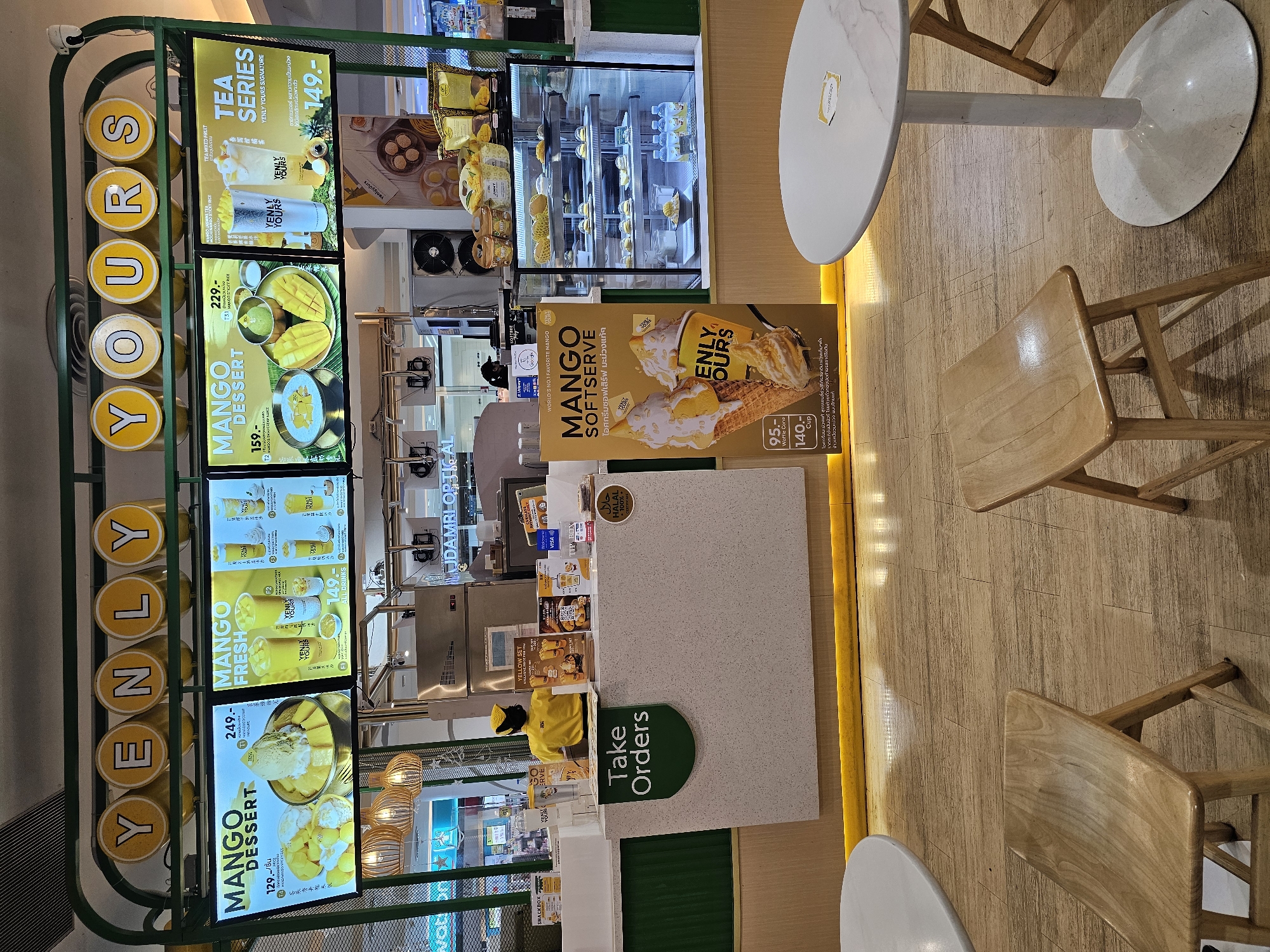 The image depicts a Yenly Yours dessert shop counter, likely located in Bangkok, Thailand, as suggested by the presence of Thai text and common mall aesthetics. The counter area is clean and modern, with a prominent digital menu board displaying various mango-themed desserts and drinks. To the left, a section labeled "Take Orders" is visible. In the center, a staff member wearing a yellow uniform and cap stands behind the counter, appearing to be interacting with customers or preparing orders. A display case to the right showcases a selection of pastries and bottled drinks. The flooring is light-colored wood laminate, and a round white table with a marble-like pattern is set up for seating. The overall impression is of a well-organized and appealing food service establishment.