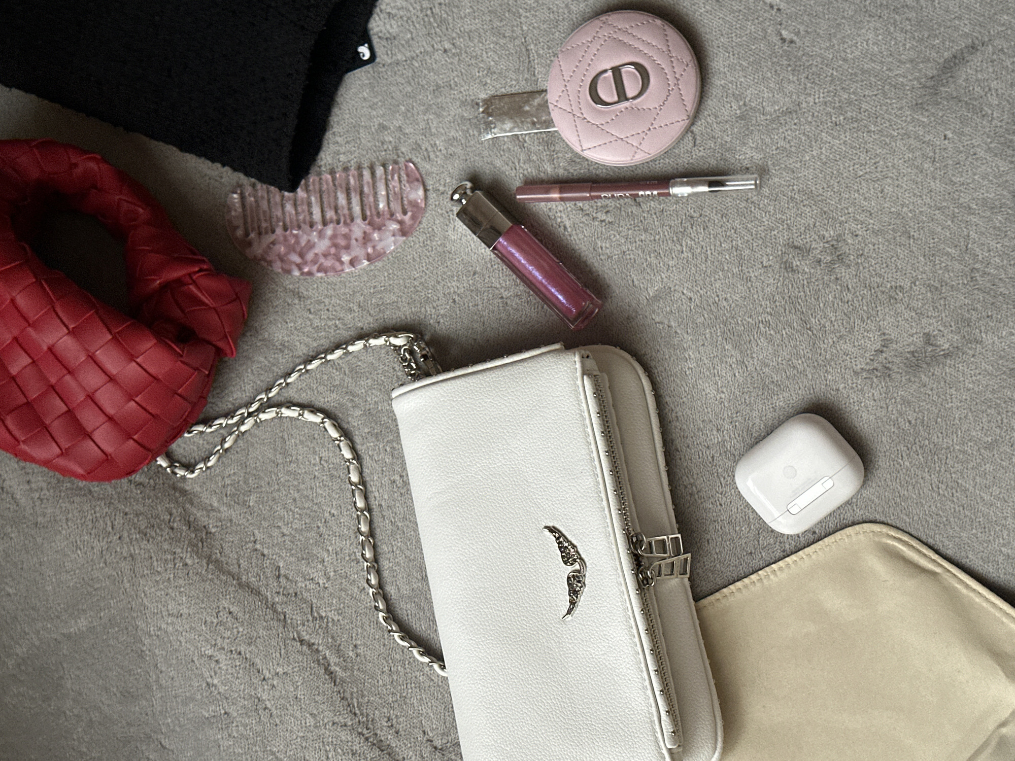 The image shows an overhead view of several personal items scattered on a gray textured surface. A small red woven bag is placed on the left. A small pink comb is located on the top left side of the image. A black fabric item rests above the red bag. A round pink Dior compact, a lip liner, and a tube of lip gloss are positioned in the upper center of the image. A white crossbody bag with a silver chain is placed diagonally across the center. To the right of the white bag, there is a white AirPods case and a tan-colored pouch. No people or elements indicating a specific location in Yerevan, Armenia, are apparent.