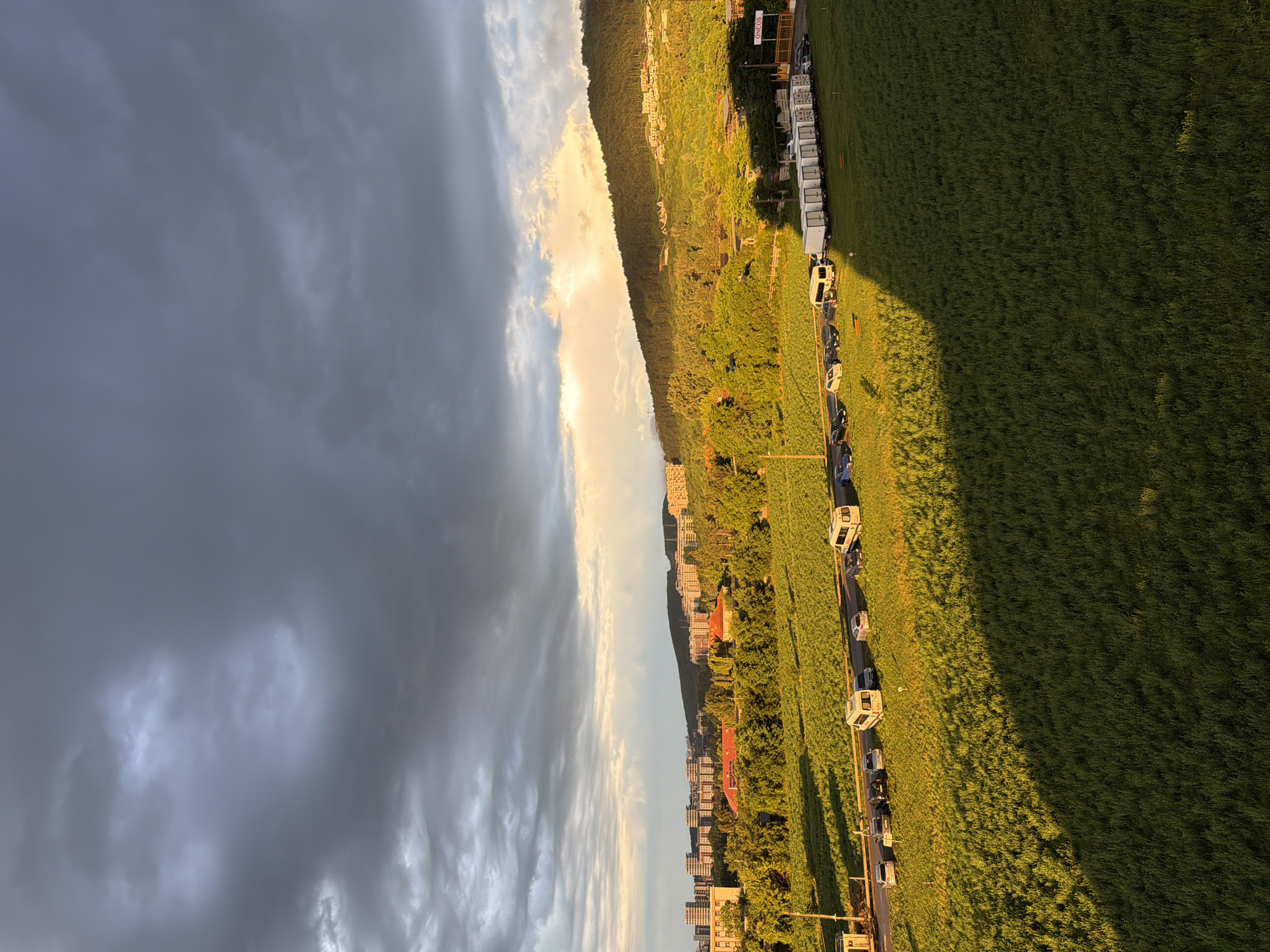 The image depicts a panoramic view of a suburban area in Florești, Romania, during the golden hour. A vast expanse of green grass, bathed in the warm glow of the setting sun, dominates the foreground, casting a long shadow across the landscape. A paved road runs horizontally across the middle ground, with a line of parked vehicles, including cars and vans, flanking it. Beyond the road, a dense cluster of green trees and vegetation stretches towards a hillside covered with more trees and buildings. In the distance, several multi-story apartment buildings are visible, interspersed with more greenery. The sky above is a dramatic canvas of dark, heavy clouds, with pockets of bright sunlight breaking through, creating a striking contrast between light and shadow. The overall scene suggests a tranquil evening in a semi-urban environment, with elements of nature and human development coexisting under a dynamic sky.