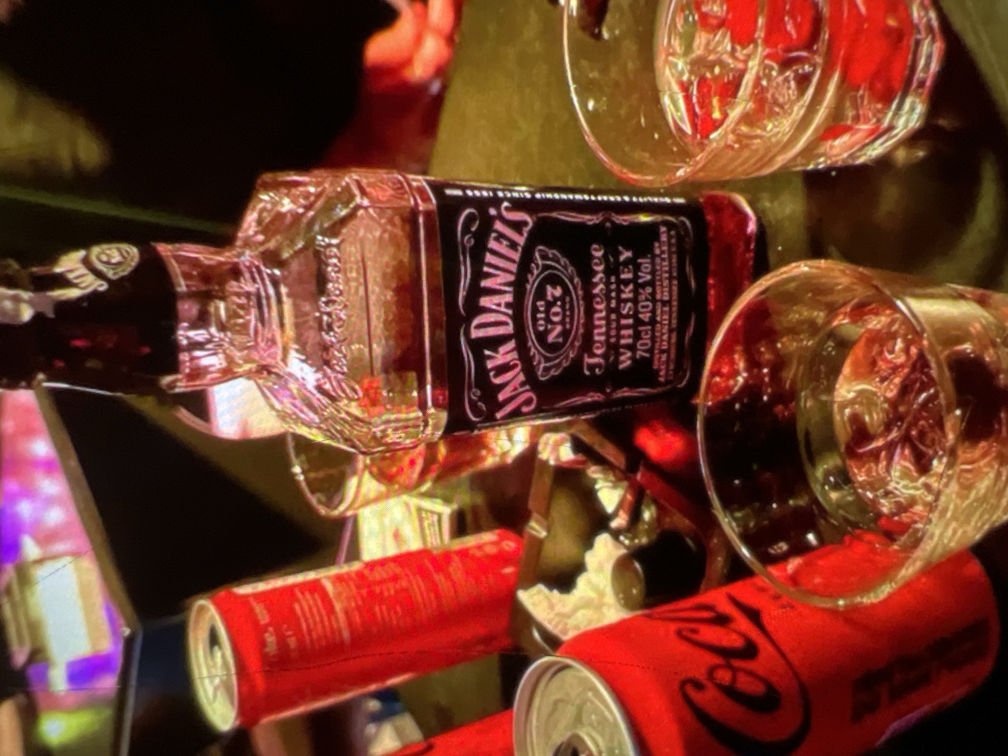The image depicts a table setting with alcoholic beverages and soft drinks, likely in a bar or restaurant. Prominently featured is a bottle of Jack Daniel's Tennessee Whiskey, alongside two glasses of whiskey on the rocks. Two cans of Coca-Cola are also visible, one fully in frame and another partially obscured. The lighting is dim, with ambient reflections suggesting a nightlife setting. There are no people visible in the image. The environment appears to be indoors. No specific details about the city of Lagos, Nigeria are discernable from the image itself.