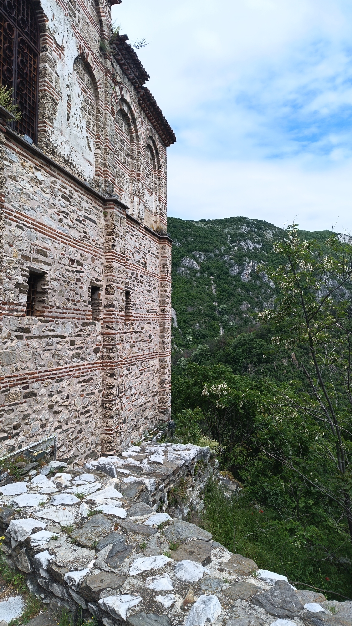 A tall, historical stone building is located in Asenovgrad, Bulgaria. The building is constructed of rough-hewn stones and red brick. It has a row of arched windows near the top, one of which has a rusty metal grate. Smaller rectangular windows are visible on the lower level of the building. The building appears old and has weathered exterior surfaces.
A stone wall built with a similar style of rocks is situated beside the building. Vegetation, including trees and shrubs, grows beside the wall. In the background, a lush green mountain is visible under a partially cloudy sky.