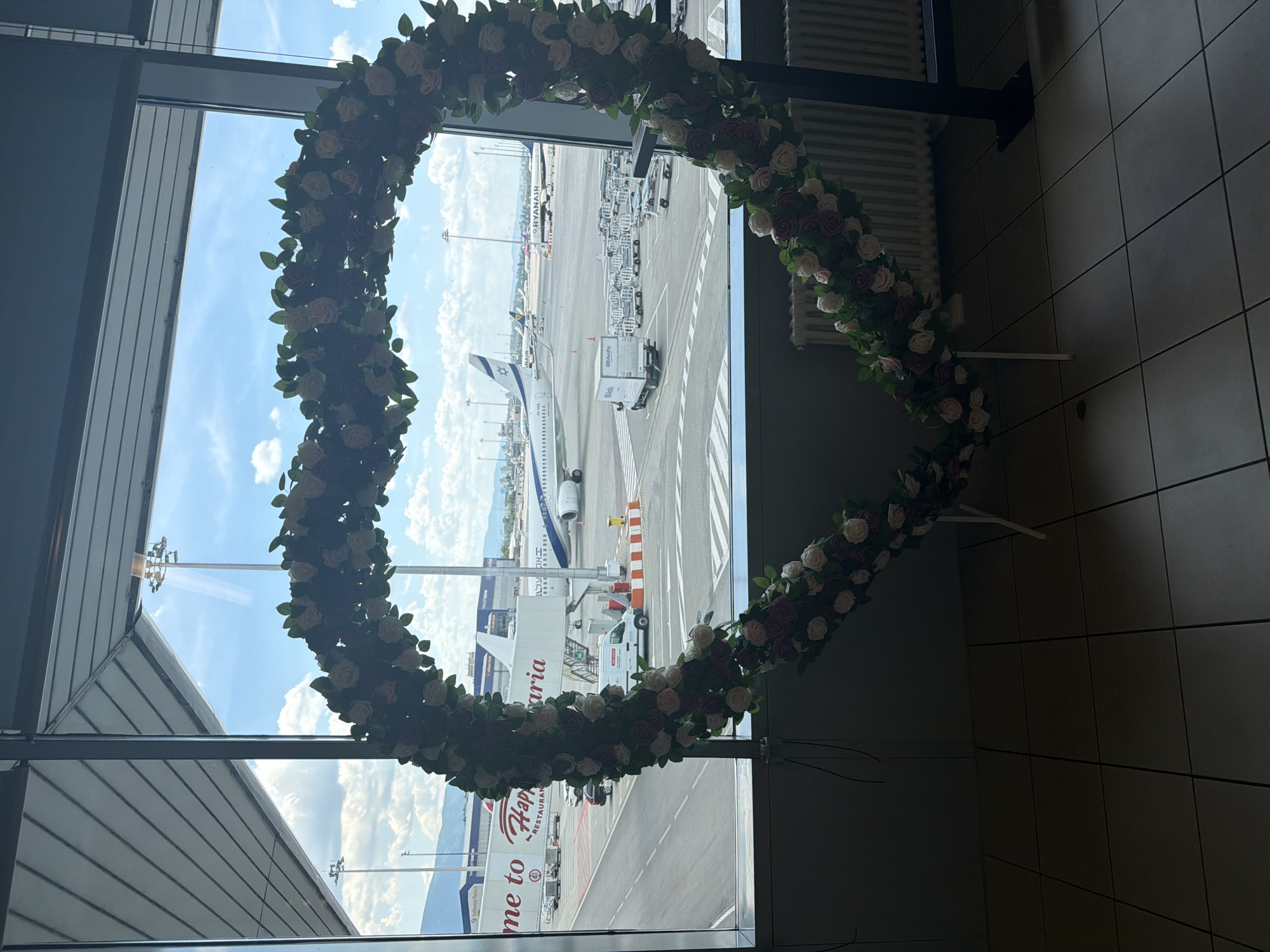 This image captures a heart-shaped floral decoration placed in front of a large window overlooking an airport tarmac. The decoration is made of artificial flowers, primarily in shades of pink, white, and dark purple, interspersed with green leaves. Through the window, several airplanes are visible, including one with "Ryanair" branding. A jet bridge is connected to another aircraft, and various airport vehicles like baggage carts and a white van are present on the tarmac. The scene appears to be set inside a terminal building, with a tiled floor and a radiator visible in the foreground. The overall impression is of a departure or arrival area with a festive or romantic decorative element.
The airport visible in the background is Sofia Airport (SOF), located in the city of Sofia, Bulgaria. The jet bridge has text that reads "Welcome to Happy Bulgaria" and "RESTAURANT," indicating a specific establishment within the airport. The presence of the "Ryanair" aircraft confirms this is a major international airport serving various airlines. The time of day is likely daytime, given the bright natural light filtering through the window and the presence of clouds in the sky. No people are clearly visible in the image.