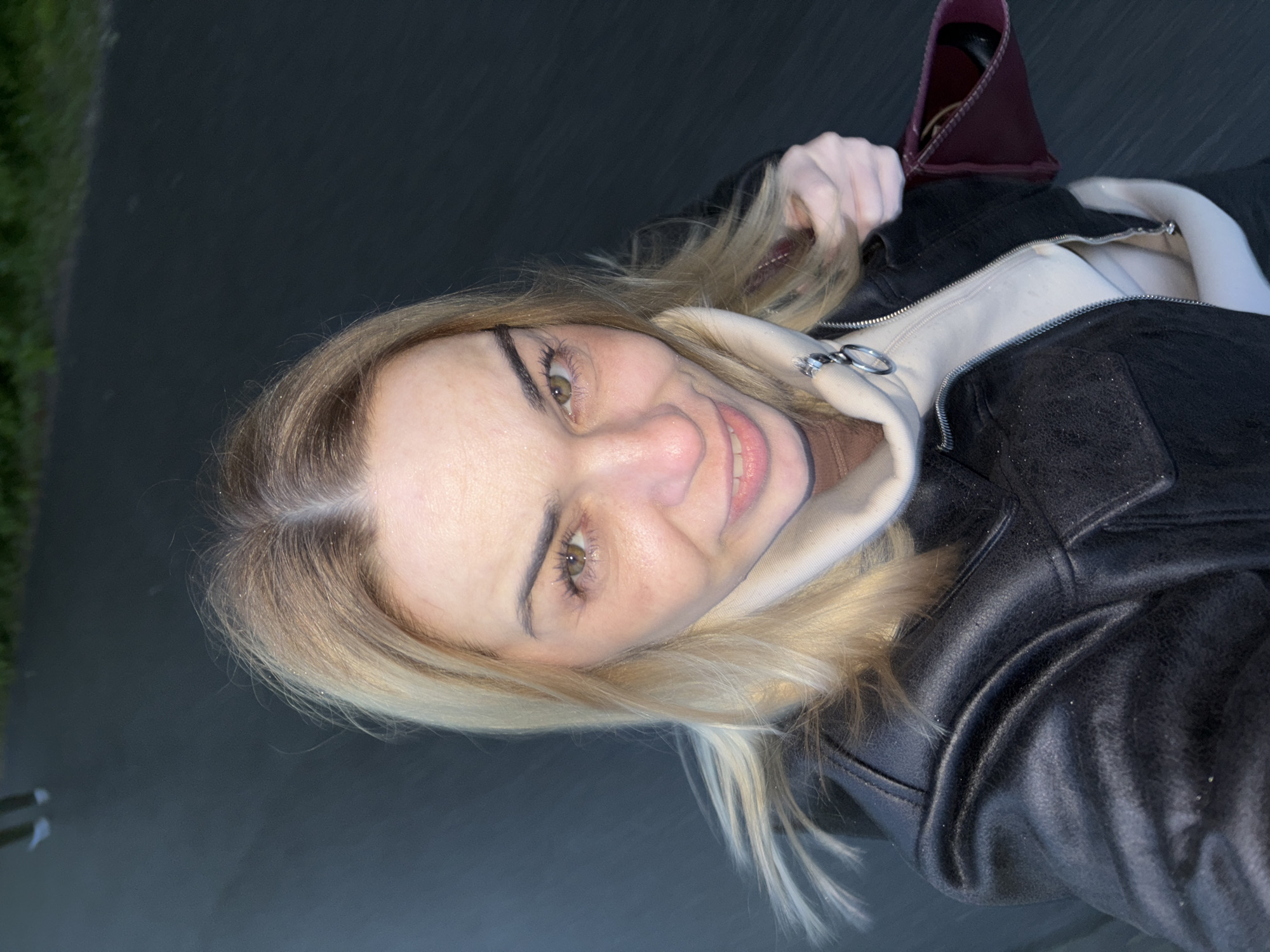 A woman with blond hair is smiling at the camera. She is wearing a black leather jacket over a light-colored hooded sweatshirt. Part of a red bag is visible in the upper right corner. The background appears to be an asphalt surface. The location appears to be a city.