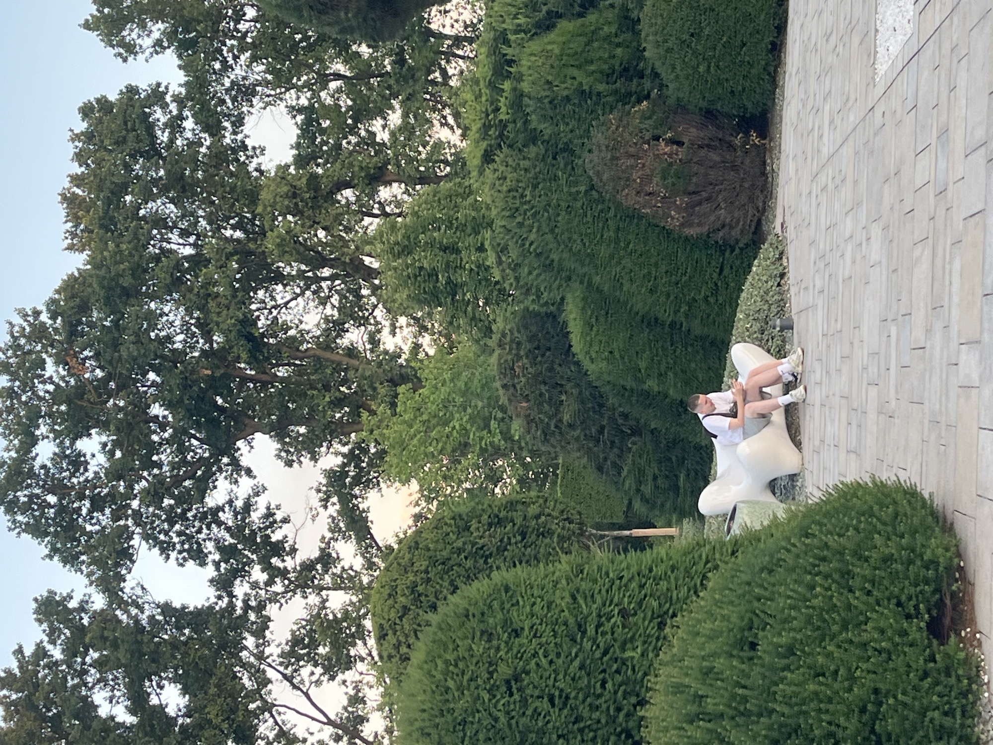 A young man sits on a white, amorphous bench in a park, looking at his phone. He is wearing a white t-shirt, grey shorts, and sneakers. The bench is surrounded by manicured evergreen bushes and trees. The ground in front of the bench is paved with light-colored rectangular tiles. In the background, large trees with dense foliage are visible against a pale sky. The image appears to be taken in a well-maintained, outdoor public space, possibly a garden or park in Krasnodar, Russia.