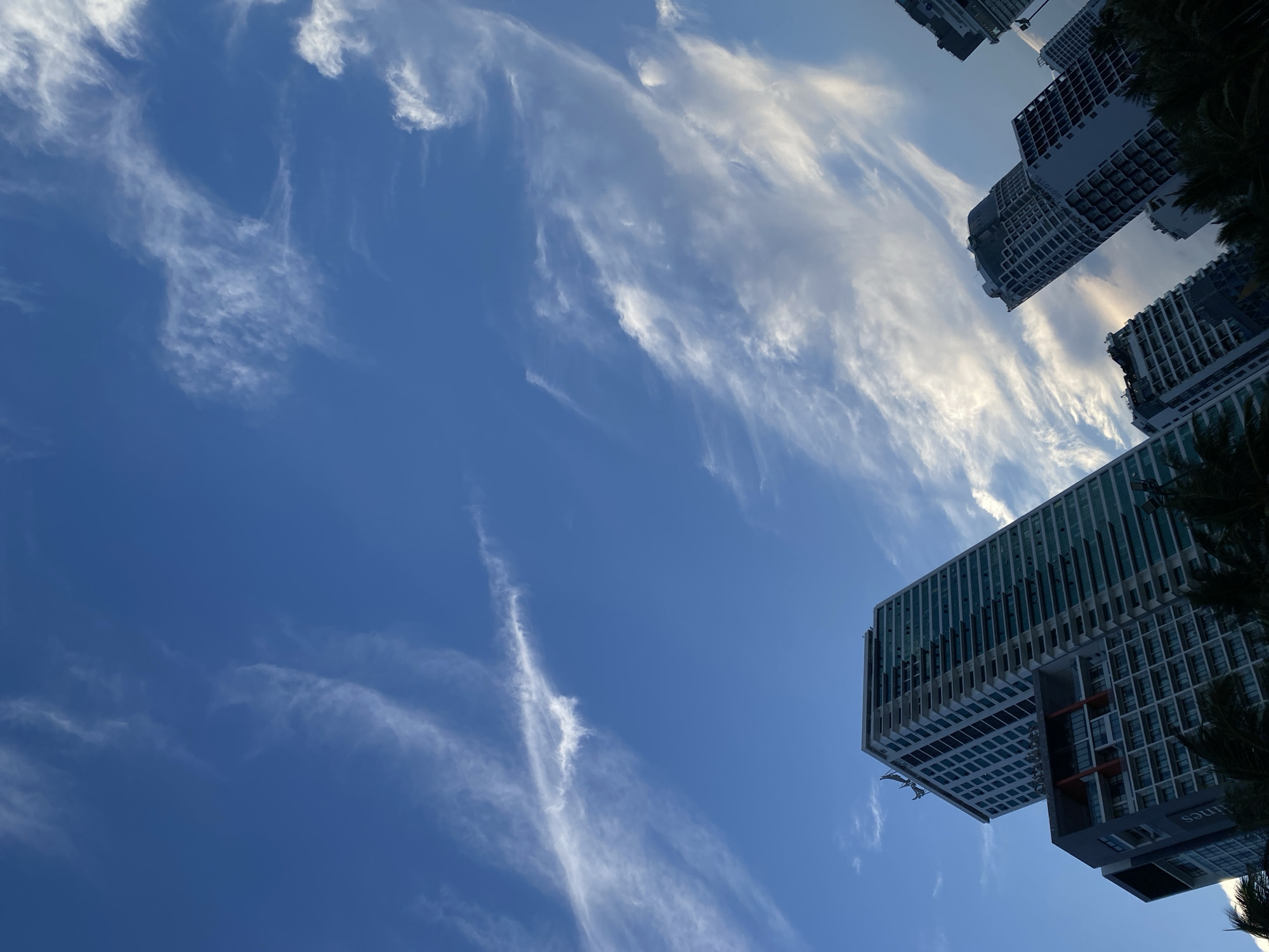 This media file shows a low-angle shot of several modern high-rise buildings against a bright blue sky with wispy white clouds. The buildings are tall and feature numerous windows, suggesting they are either residential apartments or office spaces. The perspective looking upwards emphasizes their height and modern architecture. Some greenery, possibly palm fronds, is visible on the right side, hinting at a tropical or sub-tropical location. The sky is clear and blue with thin, streaky clouds, indicative of fair weather. The sun appears to be high in the sky, suggesting it is daytime, likely late morning or early afternoon. There are no people visible in the frame, and the focus is on the architectural structures and the sky. One of the buildings has some text visible, appearing to be part of a sign or building name, but it is partially obscured and difficult to read clearly. The overall scene evokes a sense of urban development in a sunny climate. The location context provided is Nha Trang Ward, Vietnam, which aligns with the visual cues of tall buildings and tropical foliage.