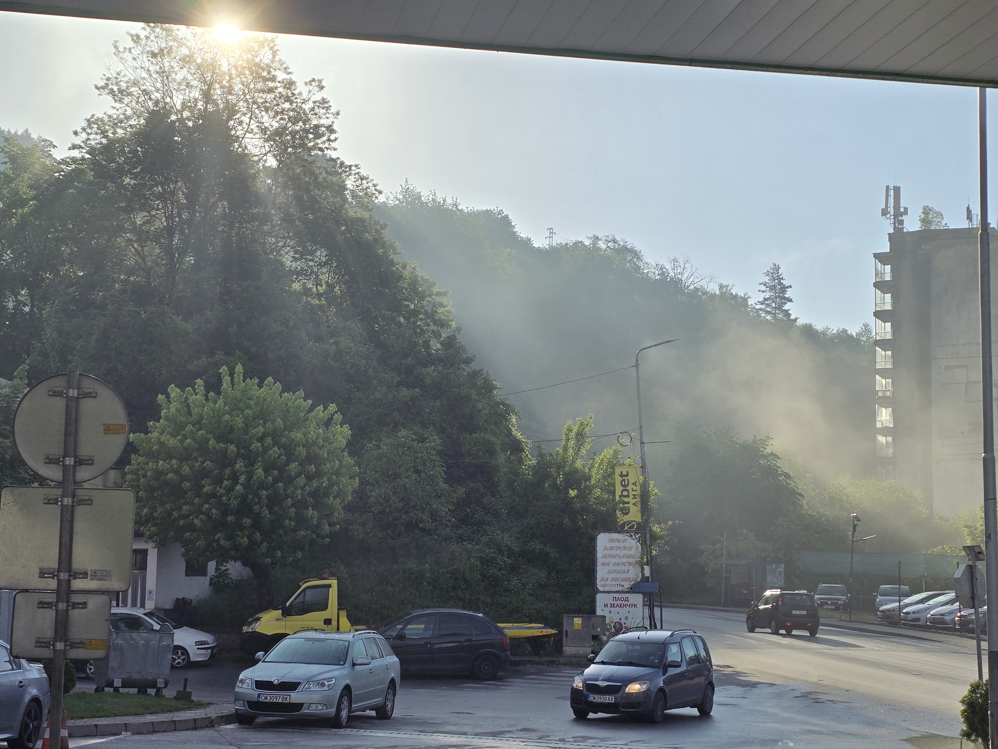 The image presents an outdoor scene depicting a street winding through a hilly, forested area, possibly a transition between urban and natural environments. The time of day appears to be either early morning or late afternoon, characterized by a bright sunburst shining through the dense canopy of a large tree on the left side of the frame. A pervasive atmospheric haze or mist blankets the scene, particularly obscuring the distant hillsides and diffusing the sunlight, creating a soft, ethereal quality.
The road, with visible wet patches or puddles, suggests recent rain or heavy morning dew. Several cars are present: a silver Skoda Octavia (license plate CM 3097 BK) is parked on the left, near a prominent circular road sign and a trash bin. Behind it, a yellow tow truck is parked among other vehicles and lush greenery. Further down the road, a dark-colored Skoda Fabia (license plate CM 3930 AX) is in motion.
On the right side of the road, a large advertising sign displays text including "erbet ЛИГА," "ЗАКУСКИ," "НА МЯСТО," "ХРАНА," "17м," and "ПЛОД И ЗЕЛЕНЧУК." In the background on the right, a tall, multi-story building with balconies and several antennas on its roof stands against the misty backdrop, partially obscured by the haze. The surrounding hills are heavily wooded, with trees appearing dark against the bright, hazy sky. No people are explicitly visible, focusing the scene on the vehicles, architecture, and natural elements.