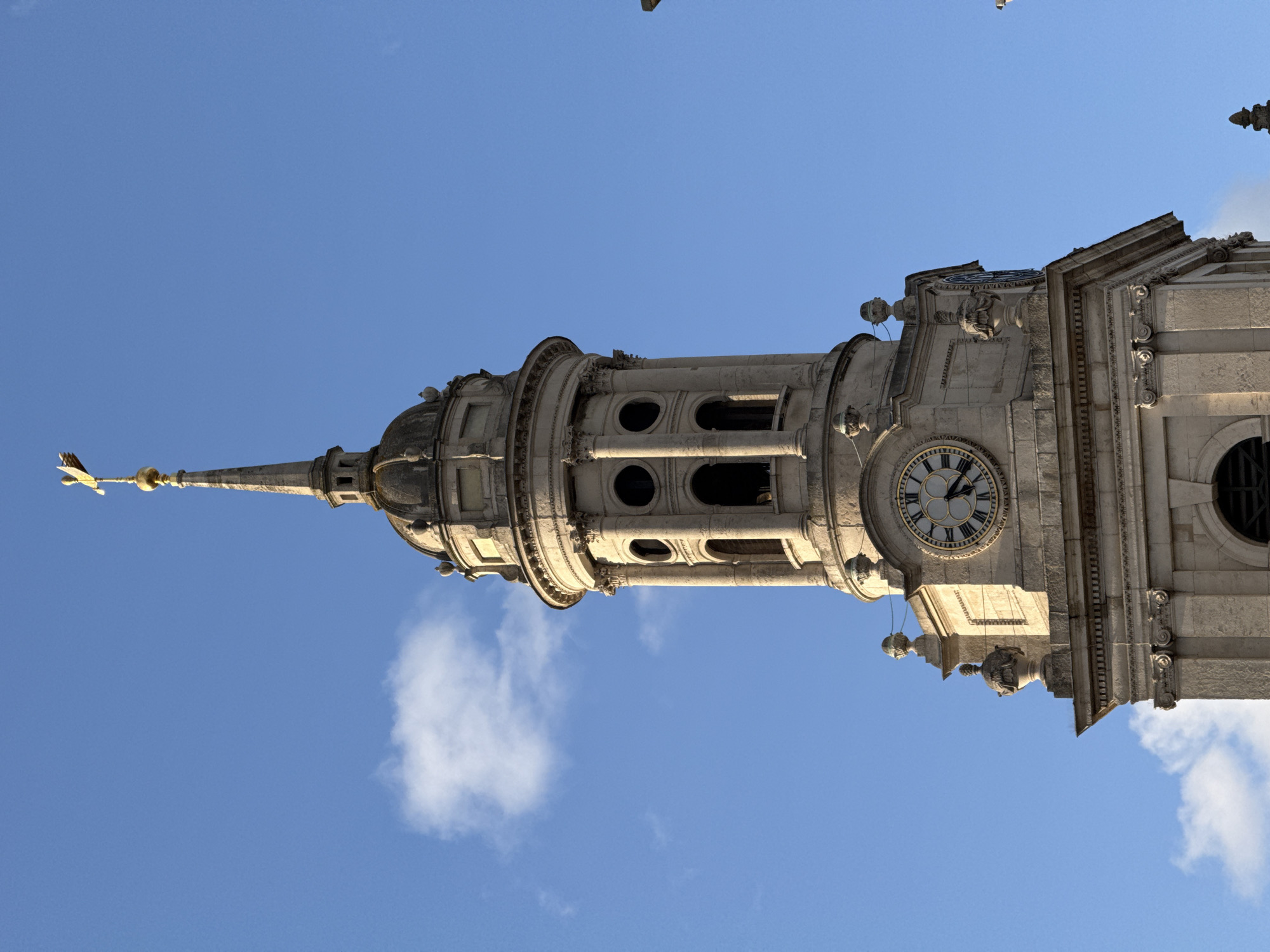 A low-angle shot shows the steeple of St. Bride's Church in London, UK, against a blue sky. The steeple features a series of diminishing tiers, culminating in a pointed spire topped with a golden weather vane. A clock face is visible on one tier. The stonework is light-colored and detailed with architectural elements. Birds are flying nearby.