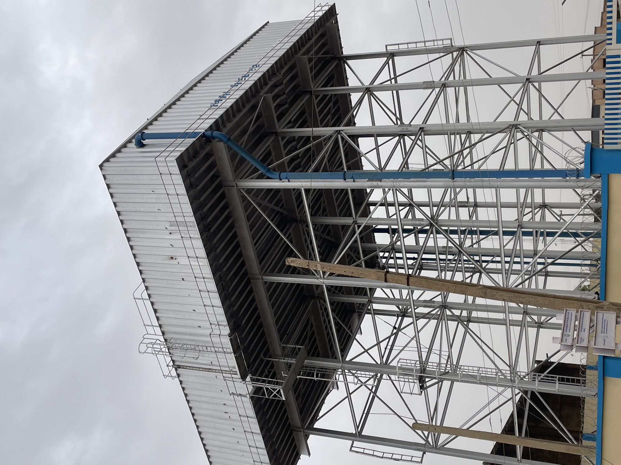 The image depicts a tall, metal water tower in Jos, Nigeria, under a cloudy sky. The tower has a silver, corrugated metal roof and is supported by a complex structure of crisscrossing silver metal beams. A blue pipe runs along the exterior of the tower. A wooden beam is temporarily attached to the metal structure. The lower section of the tower is partially obscured by a building, which has blue trim and appears to be made of concrete. Signage is visible at the base of the tower.