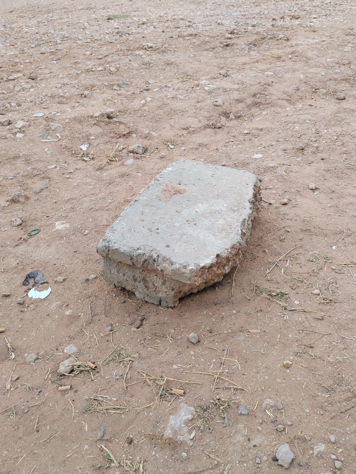 In the city of Mougheul, Algeria, a rough concrete slab rests on a dusty, uneven ground. The surface is a mix of brown earth, small pebbles, and sparse, dry grass. Scattered debris, including pieces of plastic and what appear to be cigarette butts, litters the area around the slab. The concrete itself is weathered and chipped, with visible aggregate within its structure. The lighting suggests an outdoor environment, possibly during daylight.
