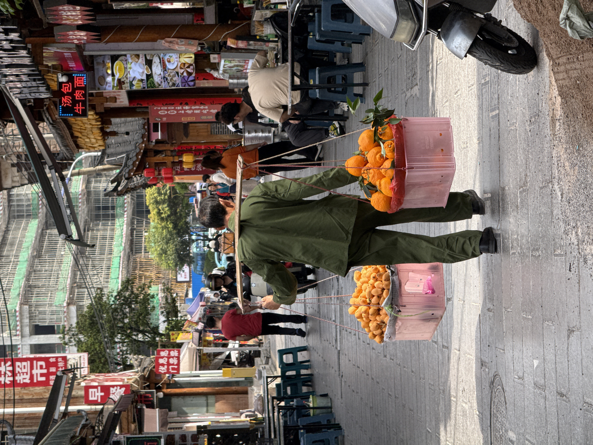 The image shows a man walking on a paved street in Zhangjiajie, China, carrying two pink plastic bins filled with oranges on a shoulder pole. He wears an olive-green jacket and pants, and dark shoes. The street is lined with shops and food stalls with signs in Chinese characters. Some people sit on small stools along the street, and a motorcycle is parked on the right. Buildings under construction with scaffolding are visible in the background.
