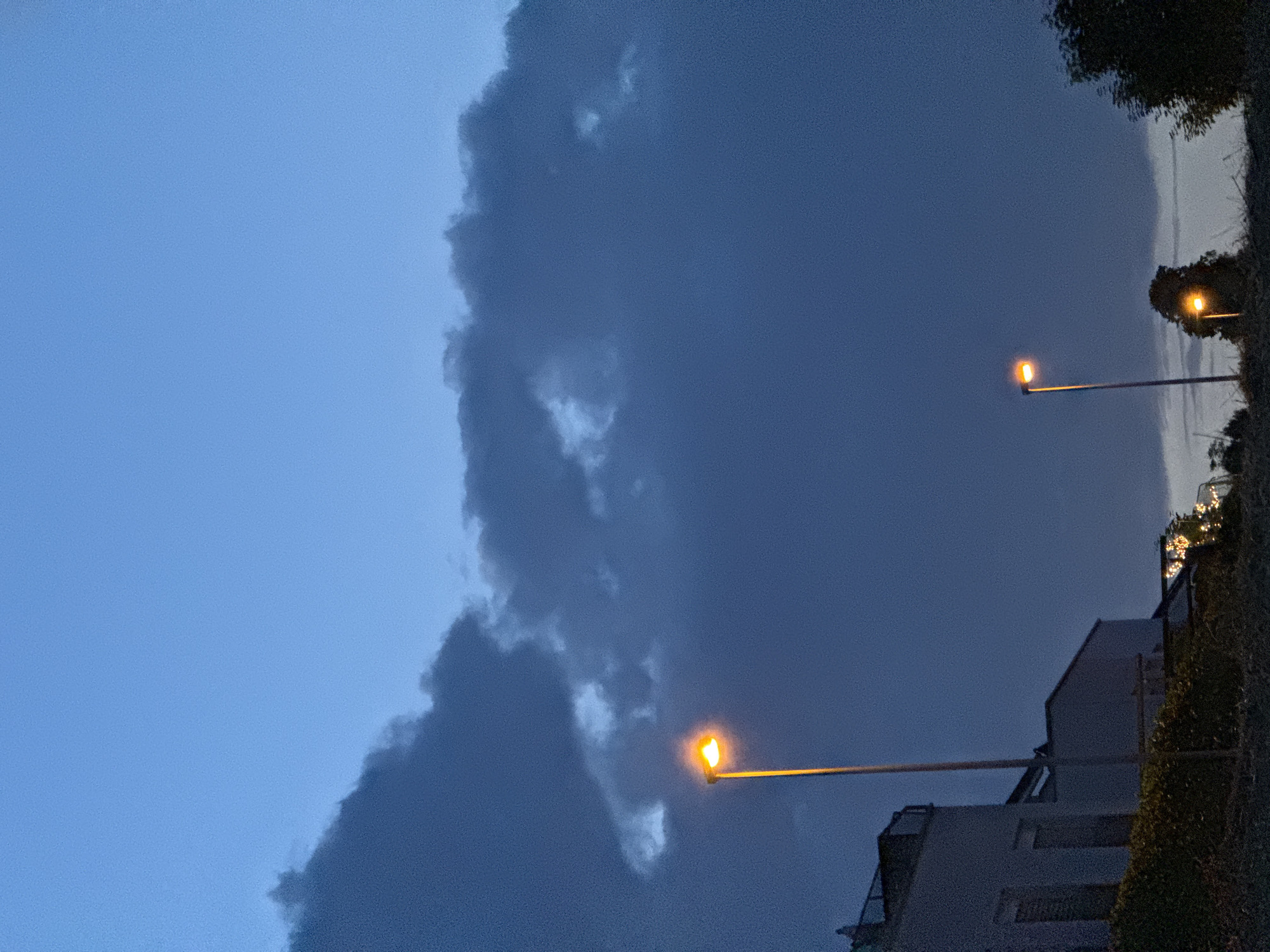 This image depicts a dimly lit outdoor scene, likely taken at dusk or night, in Lufingen, Switzerland. In the foreground, a lush green hedge runs horizontally across the bottom of the frame. Behind the hedge, a modern building with white walls and dark windows is visible on the left. Several streetlights are present. A tall, thin streetlight with a bright orange glow stands prominently in the middle left of the image. Further to the right, a shorter streetlight with a similar orange illumination is visible. The sky is a deep blue, with dark, dramatic clouds dominating the upper portion of the frame, suggesting an overcast or approaching storm. Trees, some silhouetted against the sky, are present on the far right and in the middle distance. The overall atmosphere is moody and quiet, with the artificial lights of the streetlamps providing the primary source of illumination.