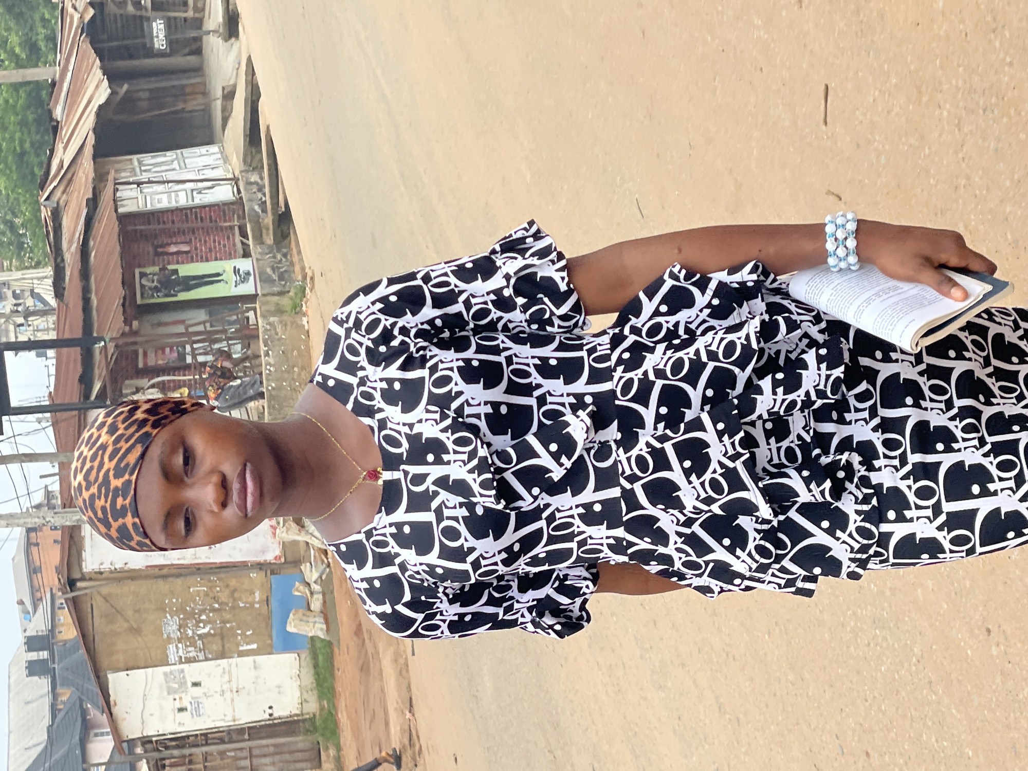 A woman stands on a dusty road in Nkpor, Nigeria. She wears a leopard-print headscarf and a black-and-white patterned dress. On her wrist, she has a beaded bracelet, and she is holding a book. The background reveals a ramshackle urban environment with buildings constructed from corrugated metal, brick, and weathered wood. A cement sign is visible on one of the buildings. The road is unpaved and has a tan hue.