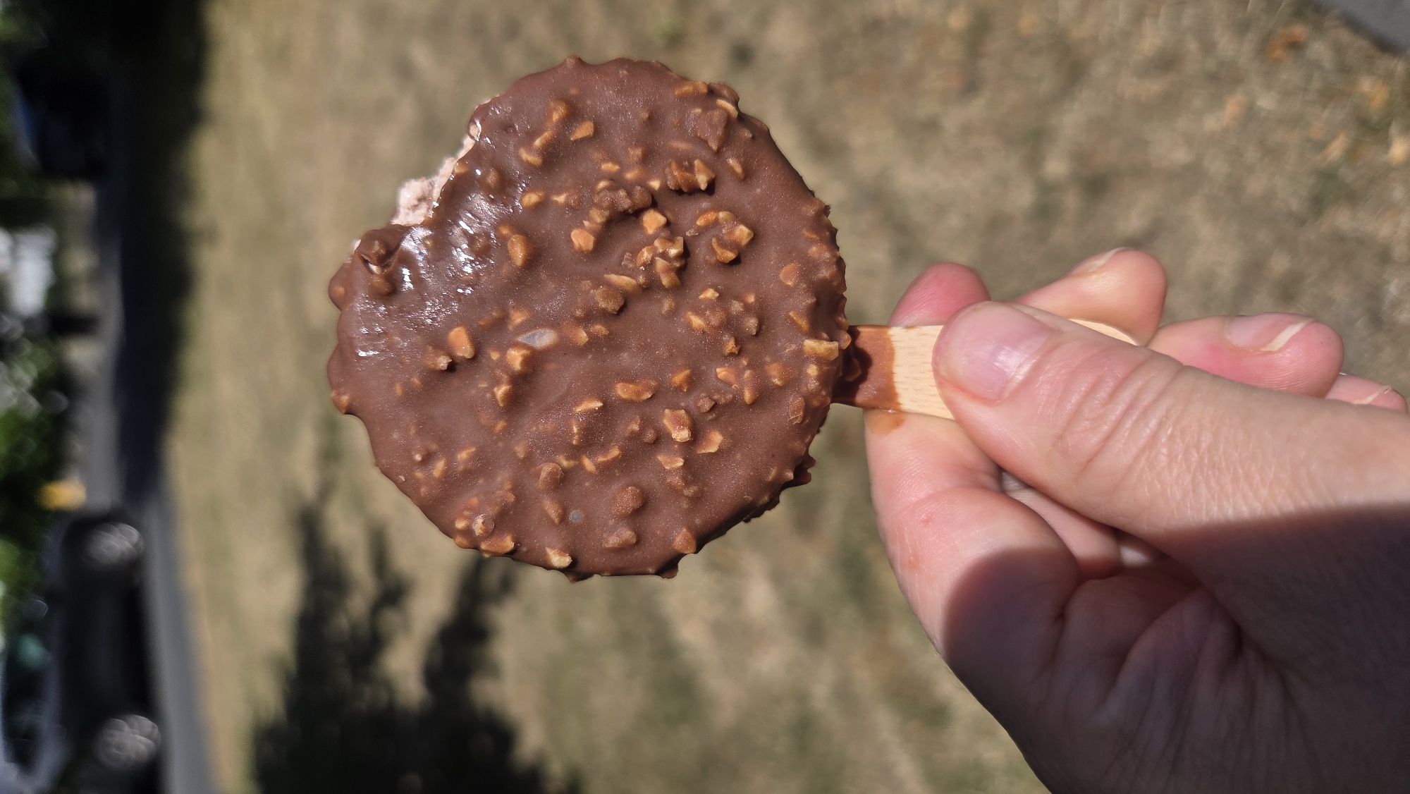 A person's hand is holding a chocolate-covered ice cream bar with nuts. The ice cream bar is circular and partially eaten. The background is blurred, showing dry grass, trees, and a road with a car. This scene appears to be in Bray, Ireland, given the context provided.